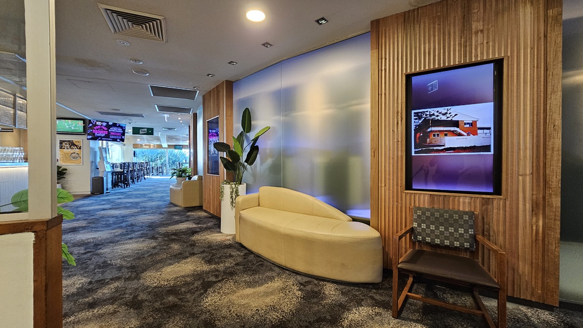 On the left, there is a hallway with a bar area visible in the distance, featuring bar stools and multiple wall-mounted TVs. In the middle, there is a frosted glass partition with a curved beige sofa and a tall potted plant in front of it. On the right, there is a wooden panelled wall with a framed digital display showing an image of a house, and a wooden chair with a patterned cushion. The floor is covered in dark, patterned carpet, and the ceiling has recessed lighting and air con vents. On the left, there is a hallway with a bar area visible in the distance, featuring bar stools and multiple wall-mounted TVs. In the middle, there is a frosted glass partition with a curved beige sofa and a tall potted plant in front of it. On the right, there is a wooden panelled wall with a framed digital display showing an image of a house, and a wooden chair with a patterned cushion. The floor is covered in dark, patterned carpet, and the ceiling has recessed lighting and air con vents.