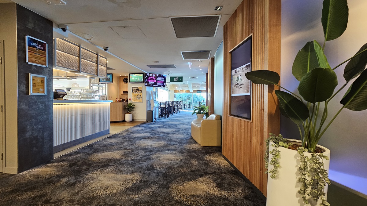 On the left, there is a bar area with a white counter, bar stools, and a menu board on the wall. In the middle, there is a hallway with a dark, patterned carpet leading to a seating area in the background, featuring tables and chairs. On the right, there is a wooden panelled wall with framed pictures, a beige sofa, and a tall potted plant. The ceiling has recessed lighting and air conditioning vents. On the left, there is a bar area with a white counter, bar stools, and a menu board on the wall. In the middle, there is a hallway with a dark, patterned carpet leading to a seating area in the background, featuring tables and chairs. On the right, there is a wooden panelled wall with framed pictures, a beige sofa, and a tall potted plant. The ceiling has recessed lighting and air conditioning vents.