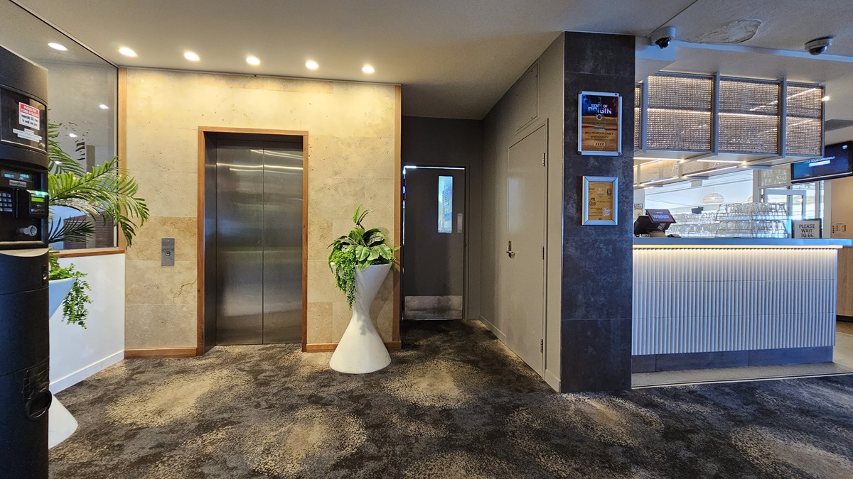 On the left, there is a black vending machine and a potted plant beside a wall. In the middle, there is a stainless steel lift with a beige wall and recessed lighting above it. In the background, there is a small hallway leading to a door. On the right, there is a reception counter with a ribbed white front and illuminated base, along with framed certificates on the wall. The floor is covered in dark, patterned carpet, and the ceiling has recessed lighting. On the left, there is a black vending machine and a potted plant beside a wall. In the middle, there is a stainless steel lift with a beige wall and recessed lighting above it. In the background, there is a small hallway leading to a door. On the right, there is a reception counter with a ribbed white front and illuminated base, along with framed certificates on the wall. The floor is covered in dark, patterned carpet, and the ceiling has recessed lighting.