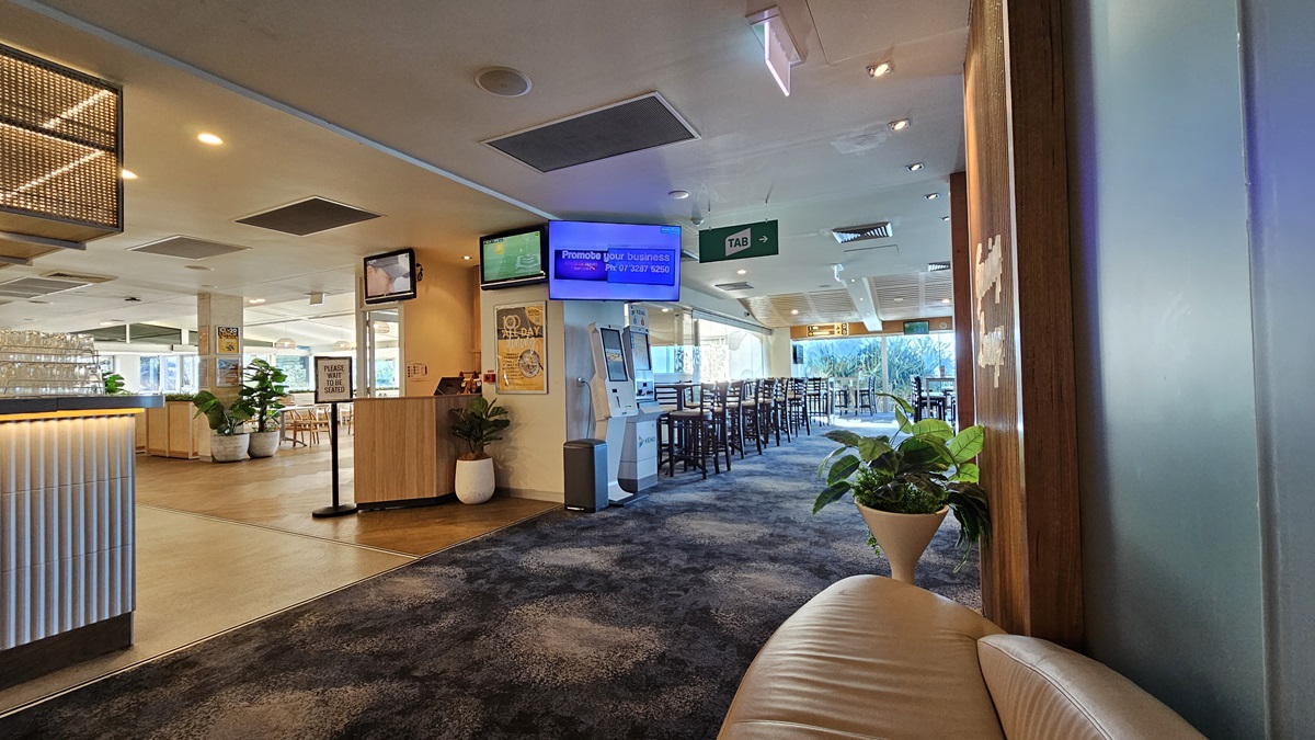 On the left, there is a reception counter with a ribbed white front and a potted plant beside it. In the middle, there is a digital kiosk and a TV screen mounted above it, with a hallway leading to a dining area in the background, featuring tables and chairs. On the right, there is a beige curved sofa and a large potted plant. The floor is covered in dark, patterned carpet, and the ceiling has recessed lighting and air conditioning vents. On the left, there is a reception counter with a ribbed white front and a potted plant beside it. In the middle, there is a digital kiosk and a TV screen mounted above it, with a hallway leading to a dining area in the background, featuring tables and chairs. On the right, there is a beige curved sofa and a large potted plant. The floor is covered in dark, patterned carpet, and the ceiling has recessed lighting and air conditioning vents.