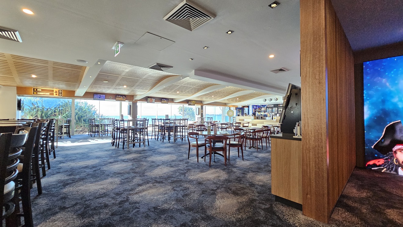 On the left, there are tall tables with high chairs and large windows offering a view of greenery. In the middle, there are multiple round tables with wooden chairs spread across the room, and a wooden ceiling with recessed lighting. In the background, there is a bar area with stools and a wall-mounted TV. On the right, there is a wooden partition wall with a digital display showing a pirate image. The floor is covered in dark, patterned carpet, and the ceiling has recessed lighting . On the left, there are tall tables with high chairs and large windows offering a view of greenery. In the middle, there are multiple round tables with wooden chairs spread across the room, and a wooden ceiling with recessed lighting. In the background, there is a bar area with stools and a wall-mounted TV. On the right, there is a wooden partition wall with a digital display showing a pirate image. The floor is covered in dark, patterned carpet, and the ceiling has recessed lighting .