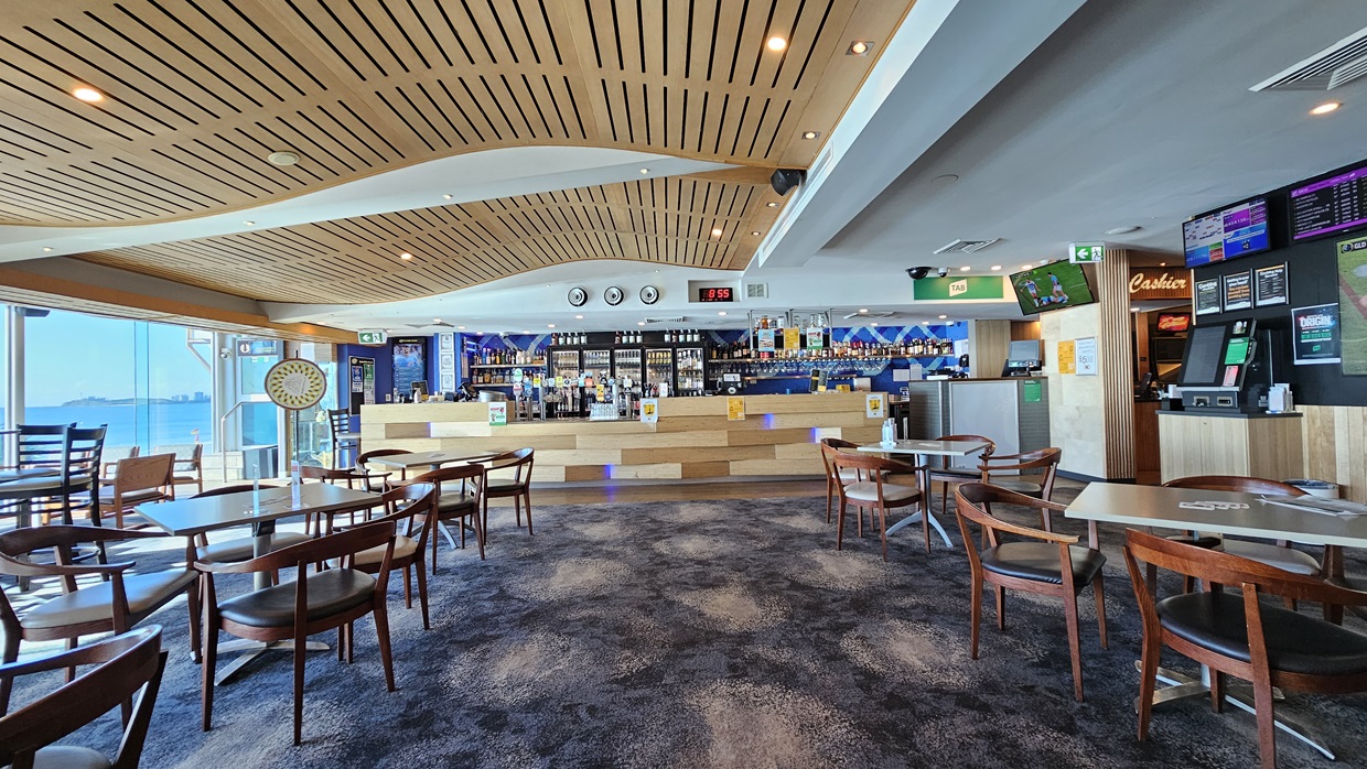 On the left, there are large windows with a view of the sea and a dartboard mounted on the wall. In the middle, there are several wooden tables and chairs on a dark, patterned carpet. In the background, there is a long bar counter with stools, various bottles, and a digital clock above it. On the right, there are more tables and chairs, a wall-mounted TV, and a gaming machine. The ceiling is wooden with a wave-like design and recessed lighting. On the left, there are large windows with a view of the sea and a dartboard mounted on the wall. In the middle, there are several wooden tables and chairs on a dark, patterned carpet. In the background, there is a long bar counter with stools, various bottles, and a digital clock above it. On the right, there are more tables and chairs, a wall-mounted TV, and a gaming machine. The ceiling is wooden with a wave-like design and recessed lighting.