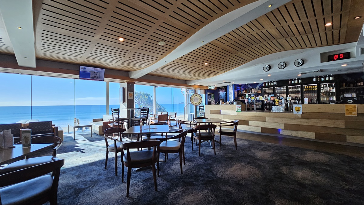 On the left, there are large windows with a view of the sea, a dartboard on the wall, and a few tables with chairs. In the middle, there are several round wooden tables with chairs on a dark, patterned carpet. In the background, there is a long bar counter with stools, various bottles, and a digital clock above it. On the right, there are more tables and chairs, a wall-mounted TV, and a gaming machine. The ceiling is wooden with a wave-like design and recessed lighting. On the left, there are large windows with a view of the sea, a dartboard on the wall, and a few tables with chairs. In the middle, there are several round wooden tables with chairs on a dark, patterned carpet. In the background, there is a long bar counter with stools, various bottles, and a digital clock above it. On the right, there are more tables and chairs, a wall-mounted TV, and a gaming machine. The ceiling is wooden with a wave-like design and recessed lighting.