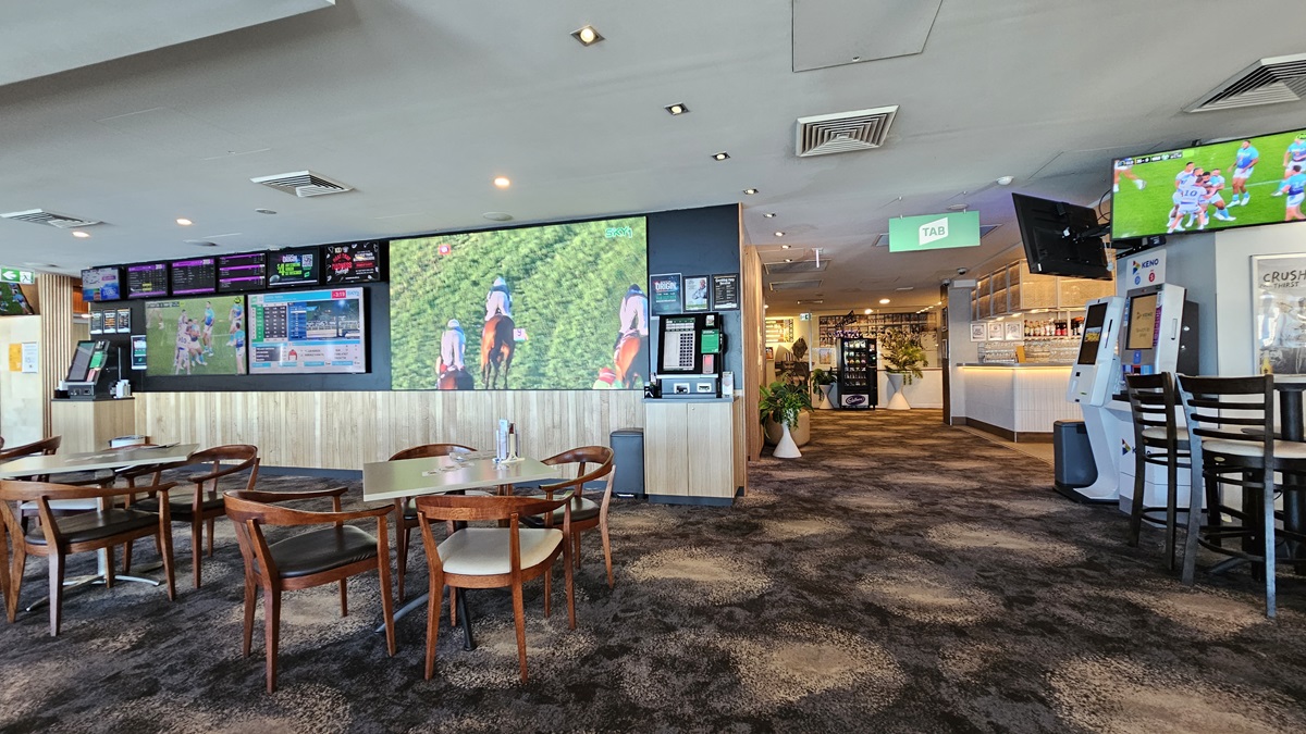On the left, there are several wooden tables with chairs and large wall-mounted screens displaying various images and information. In the middle, there is a hallway with a dark, patterned carpet leading to a bar area in the background, featuring stools and illuminated shelves with bottles. On the right, there is a gaming machine, a TV screen mounted above it, and a high table with stools. The ceiling has recessed lighting and air conditioning vents. On the left, there are several wooden tables with chairs and large wall-mounted screens displaying various images and information. In the middle, there is a hallway with a dark, patterned carpet leading to a bar area in the background, featuring stools and illuminated shelves with bottles. On the right, there is a gaming machine, a TV screen mounted above it, and a high table with stools. The ceiling has recessed lighting and air conditioning vents.