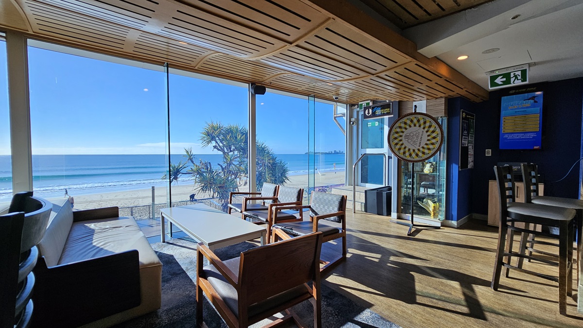 On the left, there is a black sofa with a white cushion. In the middle, there are wooden chairs and a white coffee table on a dark rug, with large windows offering a view of the beach and sea. In the background, there is a dartboard mounted on a blue wall and a vending machine. On the right, there are high tables with stools and an emergency exit sign above. The floor is wooden, and the ceiling has a wooden slat design with recessed lighting. On the left, there is a black sofa with a white cushion. In the middle, there are wooden chairs and a white coffee table on a dark rug, with large windows offering a view of the beach and sea. In the background, there is a dartboard mounted on a blue wall and a vending machine. On the right, there are high tables with stools and an emergency exit sign above. The floor is wooden, and the ceiling has a wooden slat design with recessed lighting.
