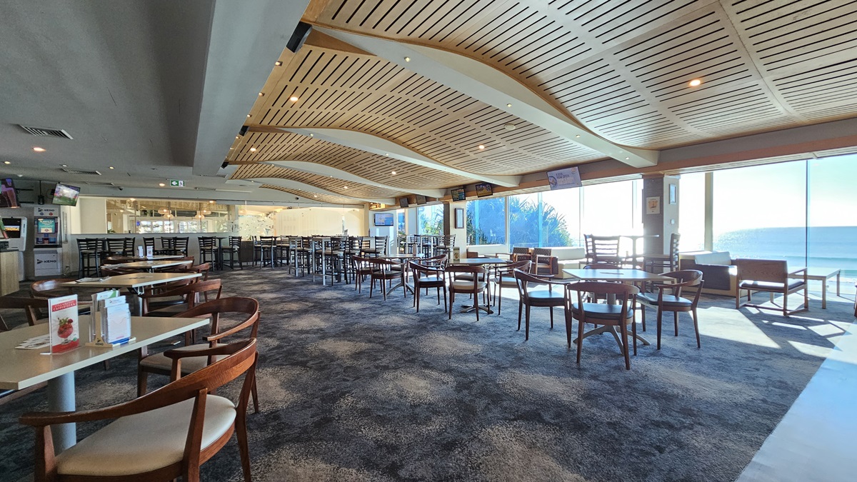 On the left, there are several wooden tables with chairs and a few wall-mounted screens. In the middle, there are more wooden tables and chairs on a dark, patterned carpet, with large windows offering a view of the sea. In the background, there is a long bar counter with stools and illuminated shelves. On the right, there are additional tables and chairs, and a lounge area with sofas. The ceiling has a wave-like wooden slat design with recessed lighting. On the left, there are several wooden tables with chairs and a few wall-mounted screens. In the middle, there are more wooden tables and chairs on a dark, patterned carpet, with large windows offering a view of the sea. In the background, there is a long bar counter with stools and illuminated shelves. On the right, there are additional tables and chairs, and a lounge area with sofas. The ceiling has a wave-like wooden slat design with recessed lighting.