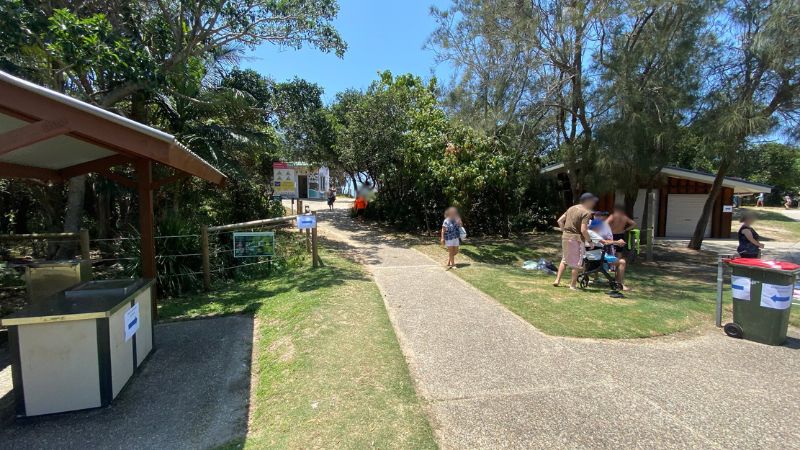 On the left, a covered barbecue area with a stone bench and a nearby fence sits next to grassy ground. On the right, a group of people stand near a small wooden building with bins on the path's edge. In the middle, a paved path leads towards a gap in the trees, revealing beach access signs and a glimpse of sandy ground in the background. The floor consists of a mix of concrete paving, grass, and patches of sand. There is no visible ceiling. On the left, a covered barbecue area with a stone bench and a nearby fence sits next to grassy ground. On the right, a group of people stand near a small wooden building with bins on the path's edge. In the middle, a paved path leads towards a gap in the trees, revealing beach access signs and a glimpse of sandy ground in the background. The floor consists of a mix of concrete paving, grass, and patches of sand. There is no visible ceiling.