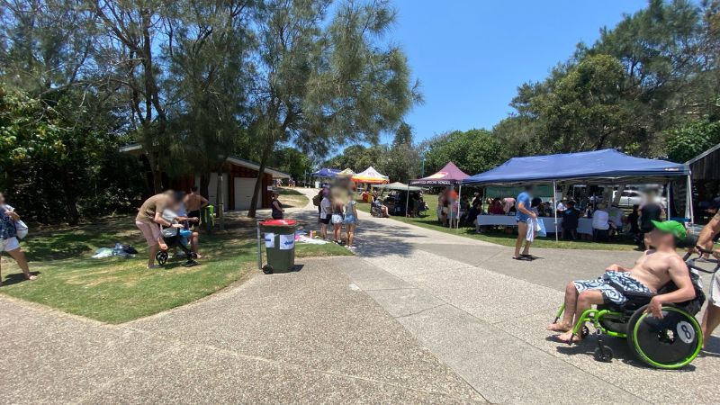 On the left, a covered barbecue area with a metal bench sits beside a railing and green grass. On the right, a group of people stand near a bin, a stroller, and a small wooden building partially visible behind trees. In the middle, a paved path leads upwards through dense greenery towards beach access signs and a glimpse of sandy ground at the path's end. The floor consists of concrete, grass, and sand. On the left, a covered barbecue area with a metal bench sits beside a railing and green grass. On the right, a group of people stand near a bin, a stroller, and a small wooden building partially visible behind trees. In the middle, a paved path leads upwards through dense greenery towards beach access signs and a glimpse of sandy ground at the path's end. The floor consists of concrete, grass, and sand.