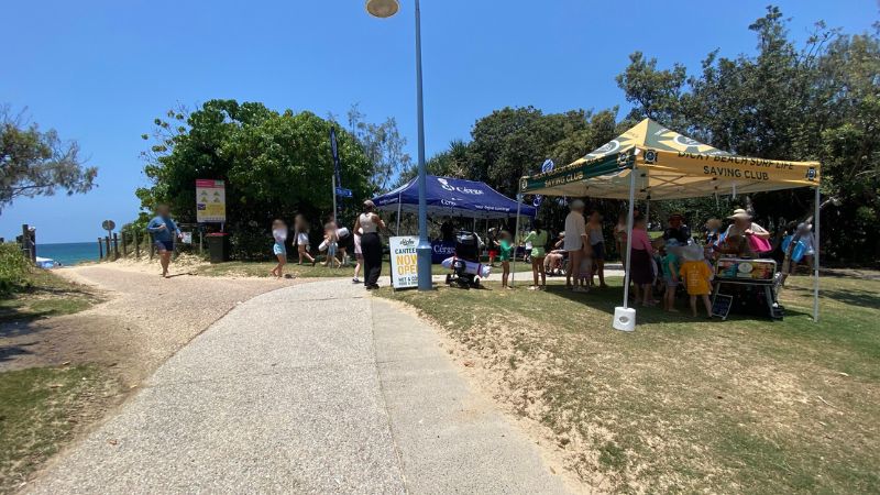 On the left, a grassy area bordered by dense trees contains a yellow warning sign and a bin near a blue "Toilets" signpost. On the right, a paved pathway curves through grass and trees, with several people walking in groups. In the middle, a dark blue gazebo branded with white text is set up on the grass, with promotional flags on either side. The foreground includes a mix of gravel and grass, with a partly visible black railing. On the left, a grassy area bordered by dense trees contains a yellow warning sign and a bin near a blue "Toilets" signpost. On the right, a paved pathway curves through grass and trees, with several people walking in groups. In the middle, a dark blue gazebo branded with white text is set up on the grass, with promotional flags on either side. The foreground includes a mix of gravel and grass, with a partly visible black railing.
