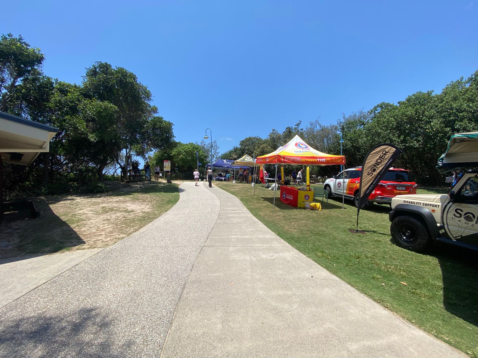 On the left, a sandy path bordered by grass and a wooden fence leads towards the beach, with a signboard and people walking nearby. On the right, two gazebos, one blue and one yellow, are set up on grass with groups of people gathered underneath. In the middle, a wide paved path runs toward the background, flanked by grass and small patches of sand. The foreground features the textured paved path and some scattered patches of dirt. A tall lamppost stands centrally. On the left, a sandy path bordered by grass and a wooden fence leads towards the beach, with a signboard and people walking nearby. On the right, two gazebos, one blue and one yellow, are set up on grass with groups of people gathered underneath. In the middle, a wide paved path runs toward the background, flanked by grass and small patches of sand. The foreground features the textured paved path and some scattered patches of dirt. A tall lamppost stands centrally.