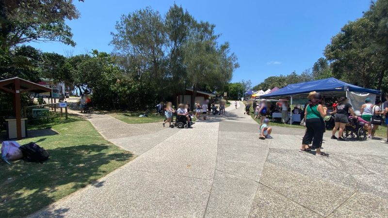 On the left, a grassy area with a small wooden shelter and a bin is surrounded by shrubs and trees. On the right, blue and white gazebos are set up on grass, with people gathered underneath and a pram nearby. In the middle, a wide paved path extends towards the background, leading to a small building and further greenery. The foreground consists of textured concrete paving, grass, and scattered shadows. On the left, a grassy area with a small wooden shelter and a bin is surrounded by shrubs and trees. On the right, blue and white gazebos are set up on grass, with people gathered underneath and a pram nearby. In the middle, a wide paved path extends towards the background, leading to a small building and further greenery. The foreground consists of textured concrete paving, grass, and scattered shadows.