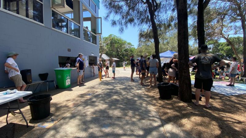 On the left, a light grey building with open service windows has people standing nearby, next to green and black bins and a white folding table. On the right, a group of people are gathered around gazebos and seated on mats under tall trees. In the middle, a paved pathway stretches forward, with scattered shadows and tree trunks along its sides. The floor consists of textured concrete, dirt, and pine straw. On the left, a light grey building with open service windows has people standing nearby, next to green and black bins and a white folding table. On the right, a group of people are gathered around gazebos and seated on mats under tall trees. In the middle, a paved pathway stretches forward, with scattered shadows and tree trunks along its sides. The floor consists of textured concrete, dirt, and pine straw.