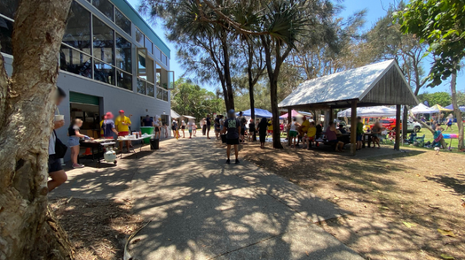 On the left, a grey building with large glass panels has people gathered beside tables holding items, with bins and vegetation nearby. On the right, a wooden pavilion with a metal roof shelters seated people, while open grass and market-style gazebos with more people are visible further back. In the middle, a paved path curves through the scene, bordered by trees casting dappled shadows. The floor includes concrete, dirt, and patches of grass. On the left, a grey building with large glass panels has people gathered beside tables holding items, with bins and vegetation nearby. On the right, a wooden pavilion with a metal roof shelters seated people, while open grass and market-style gazebos with more people are visible further back. In the middle, a paved path curves through the scene, bordered by trees casting dappled shadows. The floor includes concrete, dirt, and patches of grass.