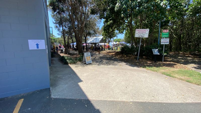 On the left, a blue-grey brick building has a white sign with a blue arrow attached to its wall. On the right, signs reading “Emergency Access” and “264” are mounted on poles near bushes, with a black information board positioned below. In the middle, a wide concrete path leads to a shaded area under trees featuring gazebos and people gathered. The foreground consists of concrete and asphalt, with greenery visible along the edges. On the left, a blue-grey brick building has a white sign with a blue arrow attached to its wall. On the right, signs reading “Emergency Access” and “264” are mounted on poles near bushes, with a black information board positioned below. In the middle, a wide concrete path leads to a shaded area under trees featuring gazebos and people gathered. The foreground consists of concrete and asphalt, with greenery visible along the edges.
