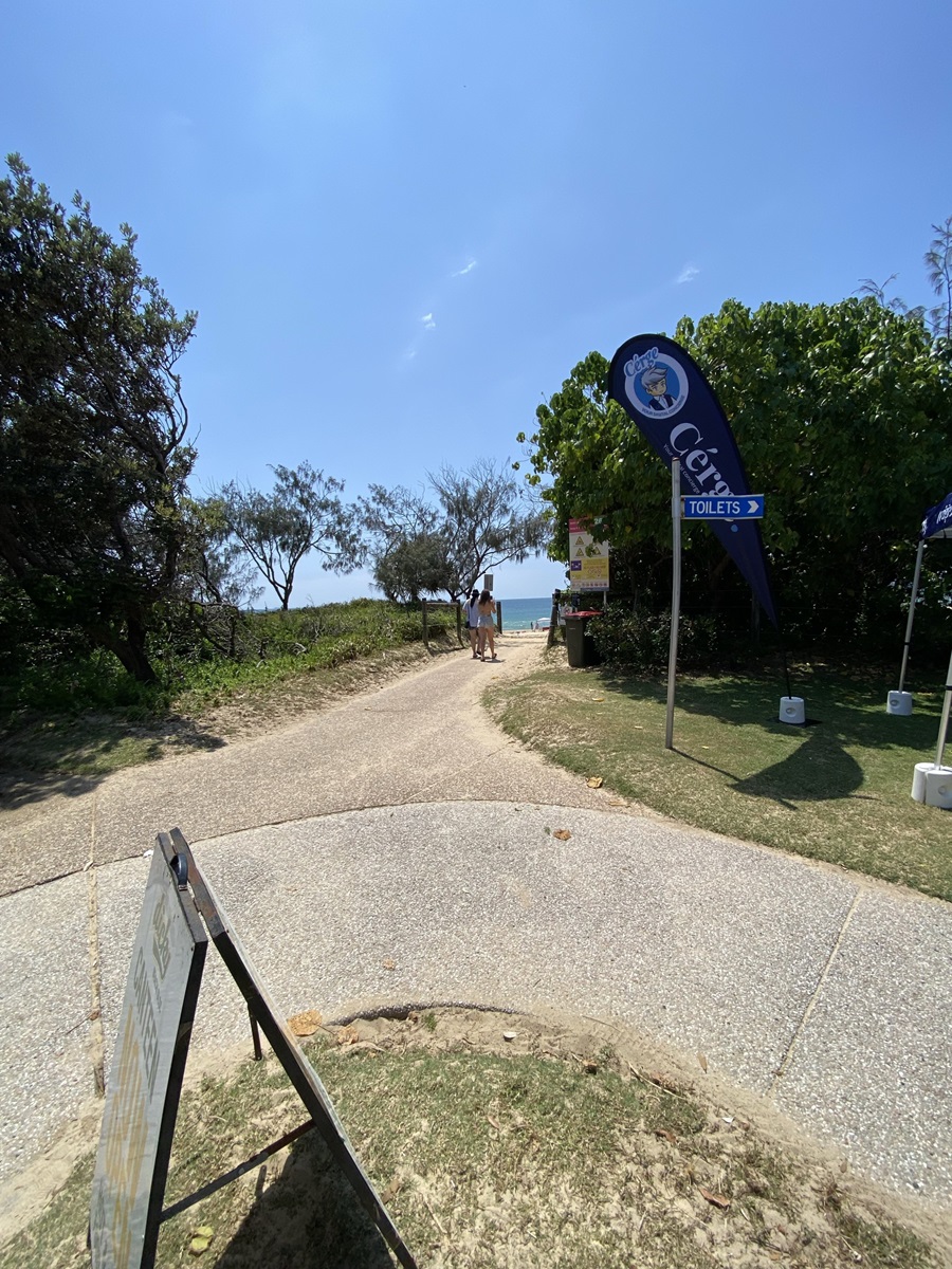 On the left, dense trees and bushes border a curved sandy path leading towards a grassy area. On the right, a blue flag marked "Toilets" stands next to a rubbish bin and lush green foliage. In the middle, a narrow, pale concrete pathway curves ahead with two people walking towards the water. The foreground consists of a section of textured concrete, trimmed grass, and part of an informational sign. On the left, dense trees and bushes border a curved sandy path leading towards a grassy area. On the right, a blue flag marked "Toilets" stands next to a rubbish bin and lush green foliage. In the middle, a narrow, pale concrete pathway curves ahead with two people walking towards the water. The foreground consists of a section of textured concrete, trimmed grass, and part of an informational sign.