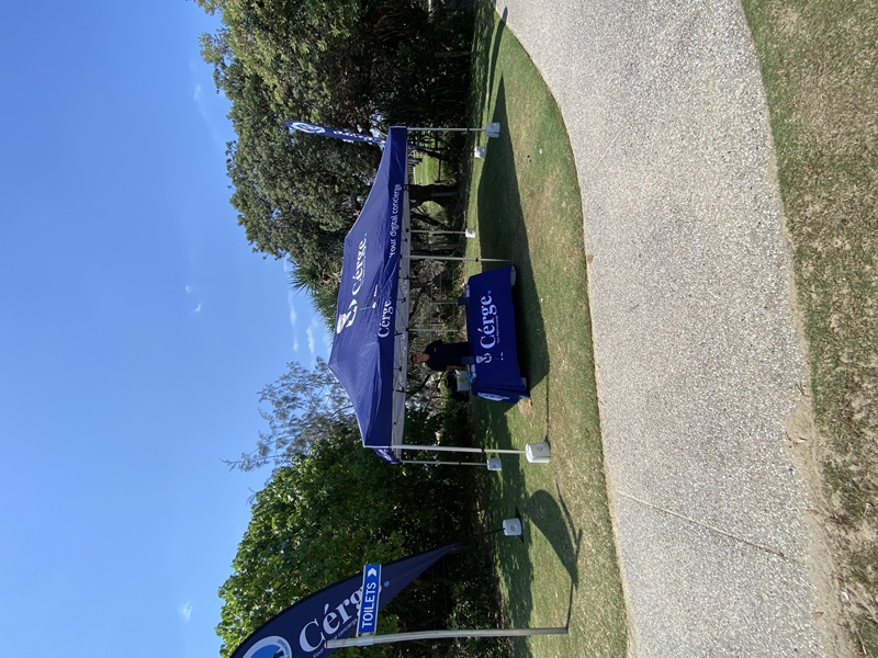 On the left, a vertical blue flag reading "Toilets" stands on grass next to bushes and trees. On the right, a cluster of dense greenery surrounds another blue flag. In the middle, a blue gazebo with "Cérge" branding on the roof and tablecloth sits on a lawn, partially shaded by trees. A person stands under the gazebo behind a table. The foreground features a textured, light-coloured curved concrete path bordered by grass. On the left, a vertical blue flag reading "Toilets" stands on grass next to bushes and trees. On the right, a cluster of dense greenery surrounds another blue flag. In the middle, a blue gazebo with "Cérge" branding on the roof and tablecloth sits on a lawn, partially shaded by trees. A person stands under the gazebo behind a table. The foreground features a textured, light-coloured curved concrete path bordered by grass.