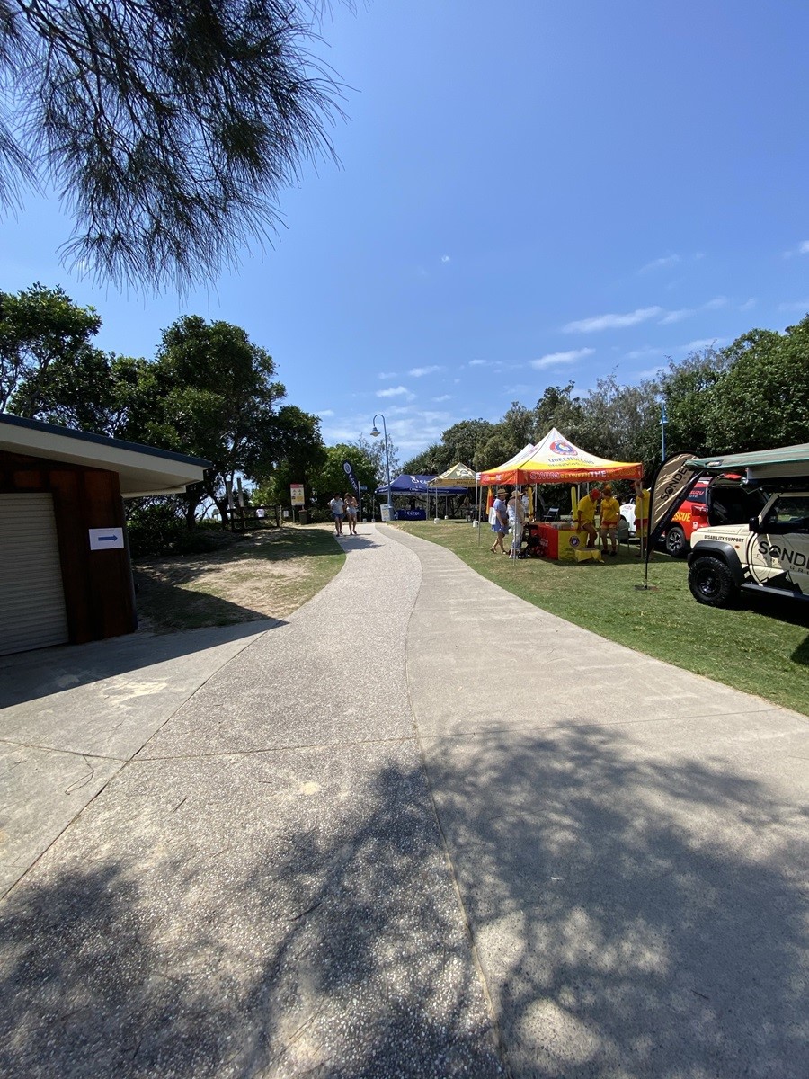 On the left, a small brown building with a closed roller door is partially in shade, next to sandy ground and grass bordered by trees. On the right, a bright yellow and orange gazebo with tables and items for display is set up on grass, alongside a parked vehicle with a partially extended awning. In the middle, a curved, textured concrete path extends into the distance, surrounded by grass and trees. The foreground includes light and shadow patterns on the concrete. On the left, a small brown building with a closed roller door is partially in shade, next to sandy ground and grass bordered by trees. On the right, a bright yellow and orange gazebo with tables and items for display is set up on grass, alongside a parked vehicle with a partially extended awning. In the middle, a curved, textured concrete path extends into the distance, surrounded by grass and trees. The foreground includes light and shadow patterns on the concrete.