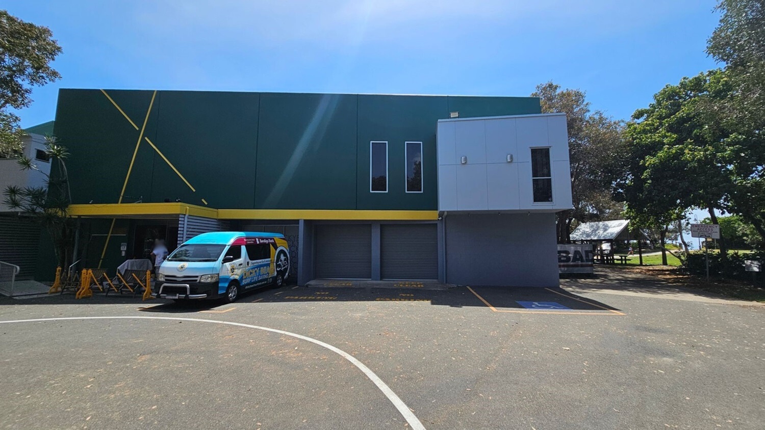 A two-storey building with a dark green wall on the left featuring yellow diagonal lines and a yellow trim above the ground level. On the right, there is a grey panelled section with a single window. At ground level, three grey roller doors are visible. In front of the left section, a white van with colourful branding is parked near bright yellow barriers. The foreground features a tarmac surface with parking markings. Trees and a small shelter are in the background on the right. A two-storey building with a dark green wall on the left featuring yellow diagonal lines and a yellow trim above the ground level. On the right, there is a grey panelled section with a single window. At ground level, three grey roller doors are visible. In front of the left section, a white van with colourful branding is parked near bright yellow barriers. The foreground features a tarmac surface with parking markings. Trees and a small shelter are in the background on the right.