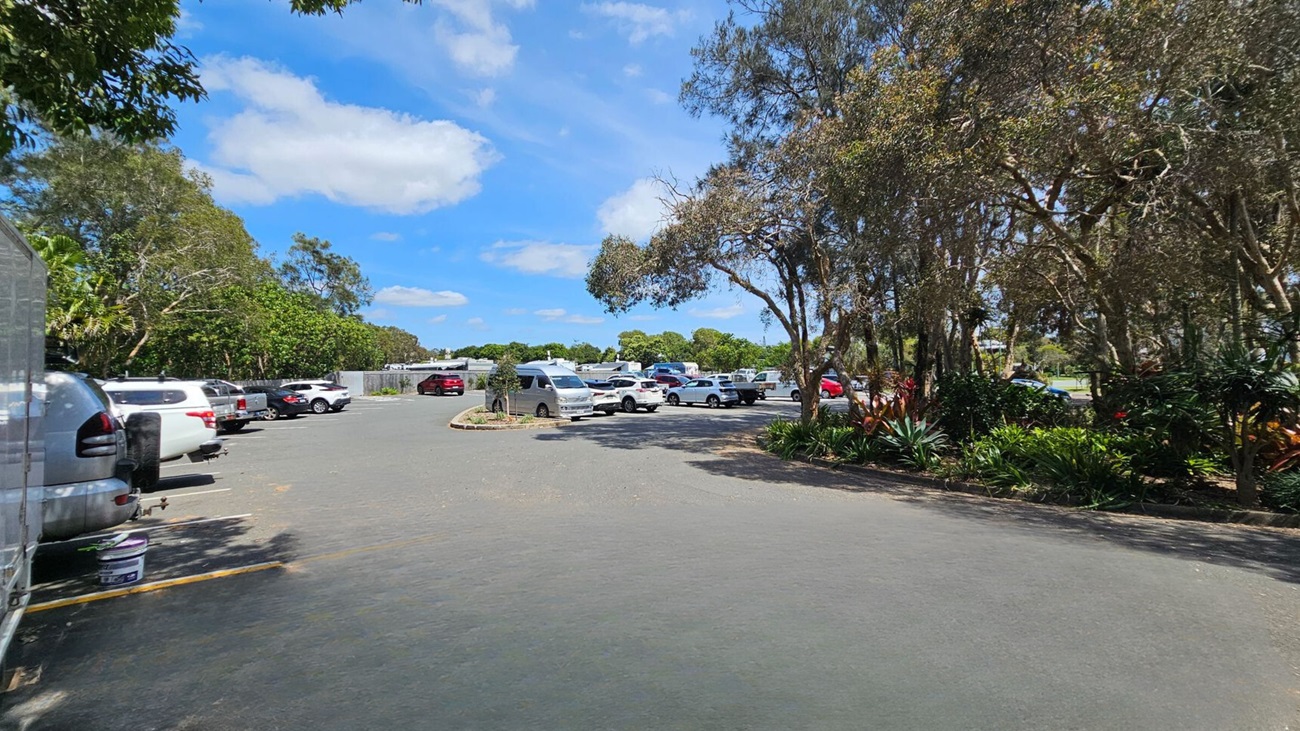 On the left, parked cars are lined along a paved parking area surrounded by green leafy trees. On the right, trees with thin trunks and thick foliage partially cover landscaped garden beds with various plants. In the middle, more parked cars, including a van, are scattered across the open paved surface. The background features more parked vehicles, fencing, and dense trees. The floor is an asphalt surface with visible wear and faint shadow patterns. On the left, parked cars are lined along a paved parking area surrounded by green leafy trees. On the right, trees with thin trunks and thick foliage partially cover landscaped garden beds with various plants. In the middle, more parked cars, including a van, are scattered across the open paved surface. The background features more parked vehicles, fencing, and dense trees. The floor is an asphalt surface with visible wear and faint shadow patterns.