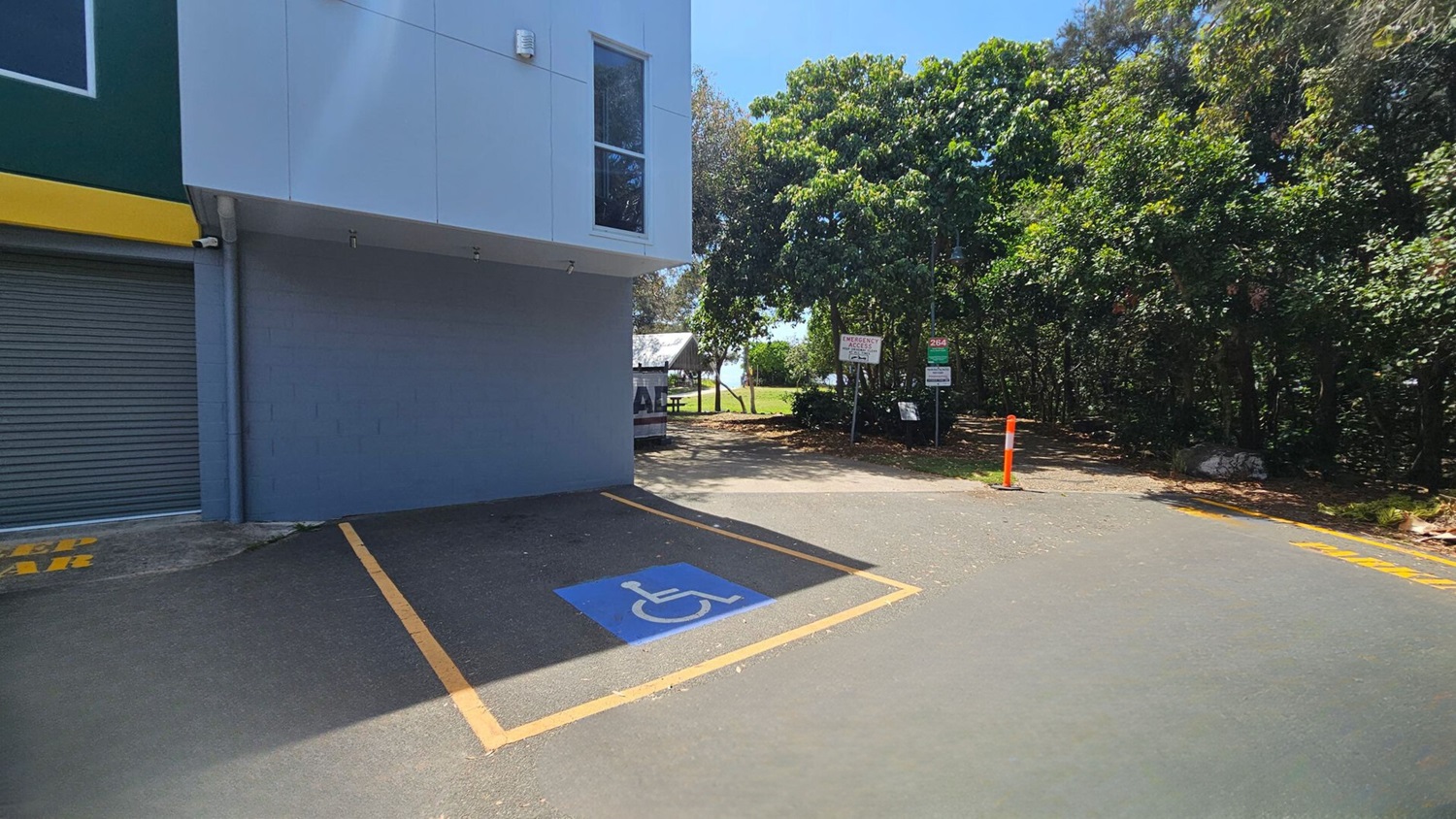 On the left, a building with a grey roller door and a yellow trim sits adjacent to a grey wall with a window above. On the right, dense green trees line the edge of the area, with signs and a bright orange bollard near a dirt pathway. In the middle, there is a marked blue disabled parking space surrounded by yellow boundary lines. The floor is asphalt, partially shaded, with a smooth, slightly worn texture. On the left, a building with a grey roller door and a yellow trim sits adjacent to a grey wall with a window above. On the right, dense green trees line the edge of the area, with signs and a bright orange bollard near a dirt pathway. In the middle, there is a marked blue disabled parking space surrounded by yellow boundary lines. The floor is asphalt, partially shaded, with a smooth, slightly worn texture.
