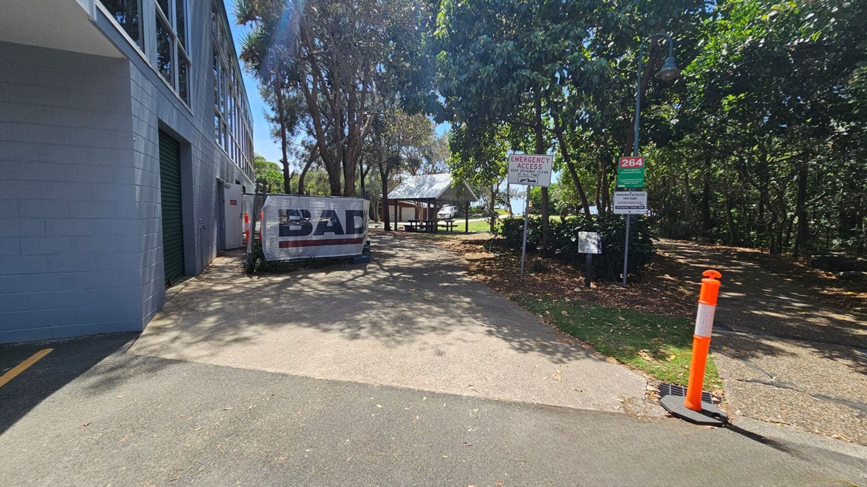 On the left, a grey building with large windows and two green roller doors extends along the edge of the image. In the middle, a concrete pathway leads to a picnic shelter with a metal roof and wooden picnic tables, surrounded by trees and grass. On the right, signs, an orange bollard, and a streetlamp are near a gravel pathway with dry leaves scattered. The floor consists of asphalt transitioning to concrete and gravel. On the left, a grey building with large windows and two green roller doors extends along the edge of the image. In the middle, a concrete pathway leads to a picnic shelter with a metal roof and wooden picnic tables, surrounded by trees and grass. On the right, signs, an orange bollard, and a streetlamp are near a gravel pathway with dry leaves scattered. The floor consists of asphalt transitioning to concrete and gravel.