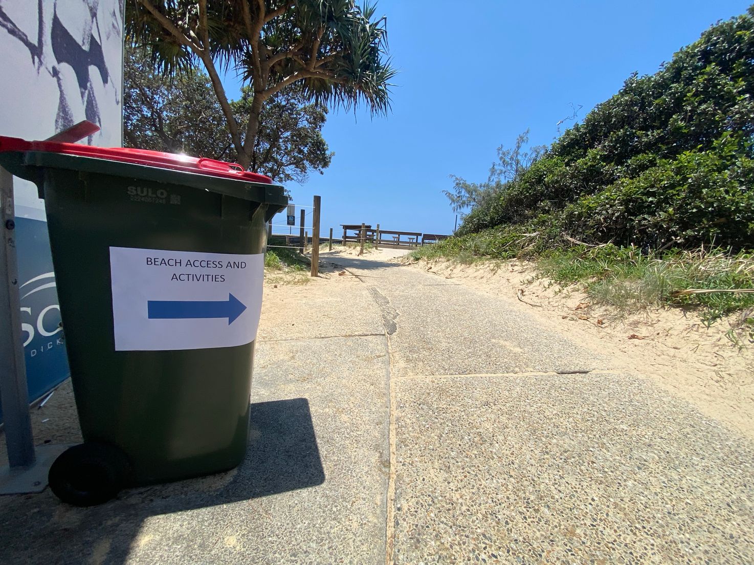 On the left, a green wheelie bin with a red lid stands beside a building, displaying a sign with "Beach Access and Activities" and an arrow pointing right. On the right, dense green shrubs and grass extend along a sandy slope. In the middle, a textured concrete path leads uphill, bordered by sand and grass, towards a wooden railing and a bench in the background. The floor transitions from concrete to sand, with patches of shadow near the bin. On the left, a green wheelie bin with a red lid stands beside a building, displaying a sign with "Beach Access and Activities" and an arrow pointing right. On the right, dense green shrubs and grass extend along a sandy slope. In the middle, a textured concrete path leads uphill, bordered by sand and grass, towards a wooden railing and a bench in the background. The floor transitions from concrete to sand, with patches of shadow near the bin.