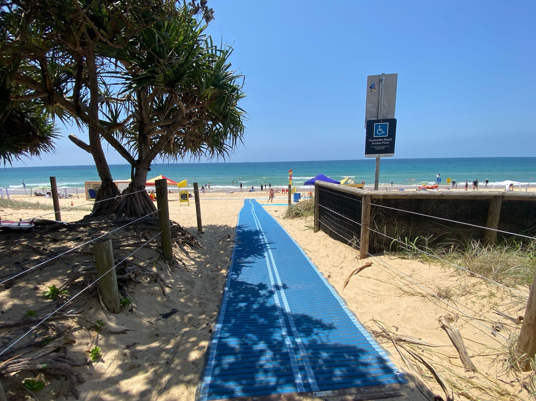 On the left, a large pandanus tree with exposed roots is surrounded by sandy ground and wooden posts connected by wire. On the right, a wooden fence with wire mesh follows the edge of the sandy pathway, with a sign for accessible beach access attached to a post. In the middle, a blue mobility mat stretches across the sand, leading towards the shoreline. The floor consists of sand with patches of vegetation near the edges of the path. On the left, a large pandanus tree with exposed roots is surrounded by sandy ground and wooden posts connected by wire. On the right, a wooden fence with wire mesh follows the edge of the sandy pathway, with a sign for accessible beach access attached to a post. In the middle, a blue mobility mat stretches across the sand, leading towards the shoreline. The floor consists of sand with patches of vegetation near the edges of the path.