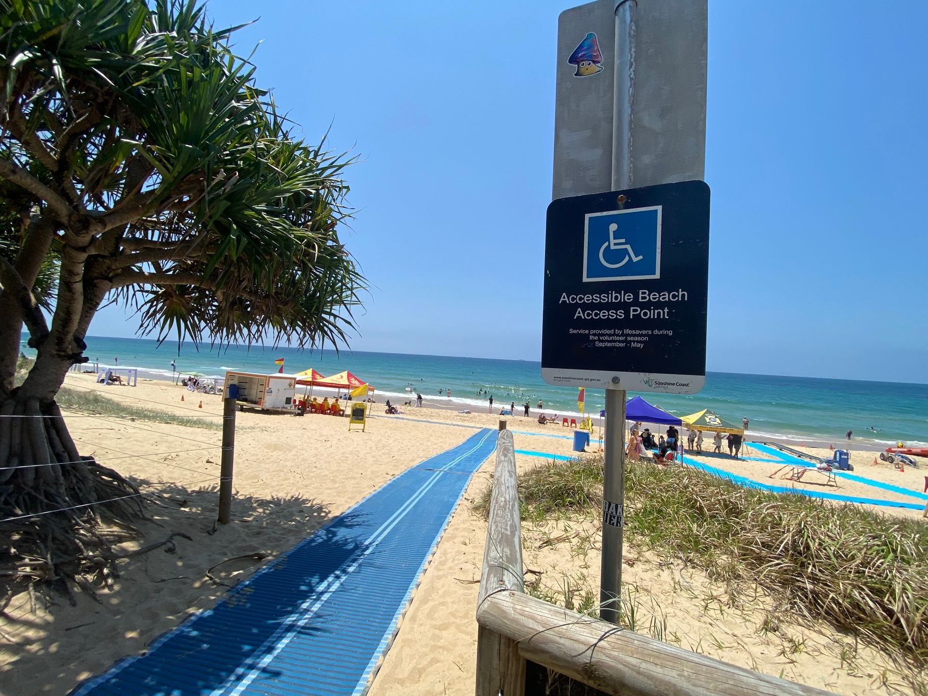On the left, a large pandanus tree with thick foliage rests on sandy ground near a wooden post with wire fencing. On the right, a blue sign on a metal post reads "Accessible Beach Access Point" above sand and grass. In the middle, a blue mobility mat stretches over the sand towards the beach, with tents and a lifeguard hut visible in the background. The floor is sandy with some grassy patches near the edges of the mat. On the left, a large pandanus tree with thick foliage rests on sandy ground near a wooden post with wire fencing. On the right, a blue sign on a metal post reads "Accessible Beach Access Point" above sand and grass. In the middle, a blue mobility mat stretches over the sand towards the beach, with tents and a lifeguard hut visible in the background. The floor is sandy with some grassy patches near the edges of the mat.