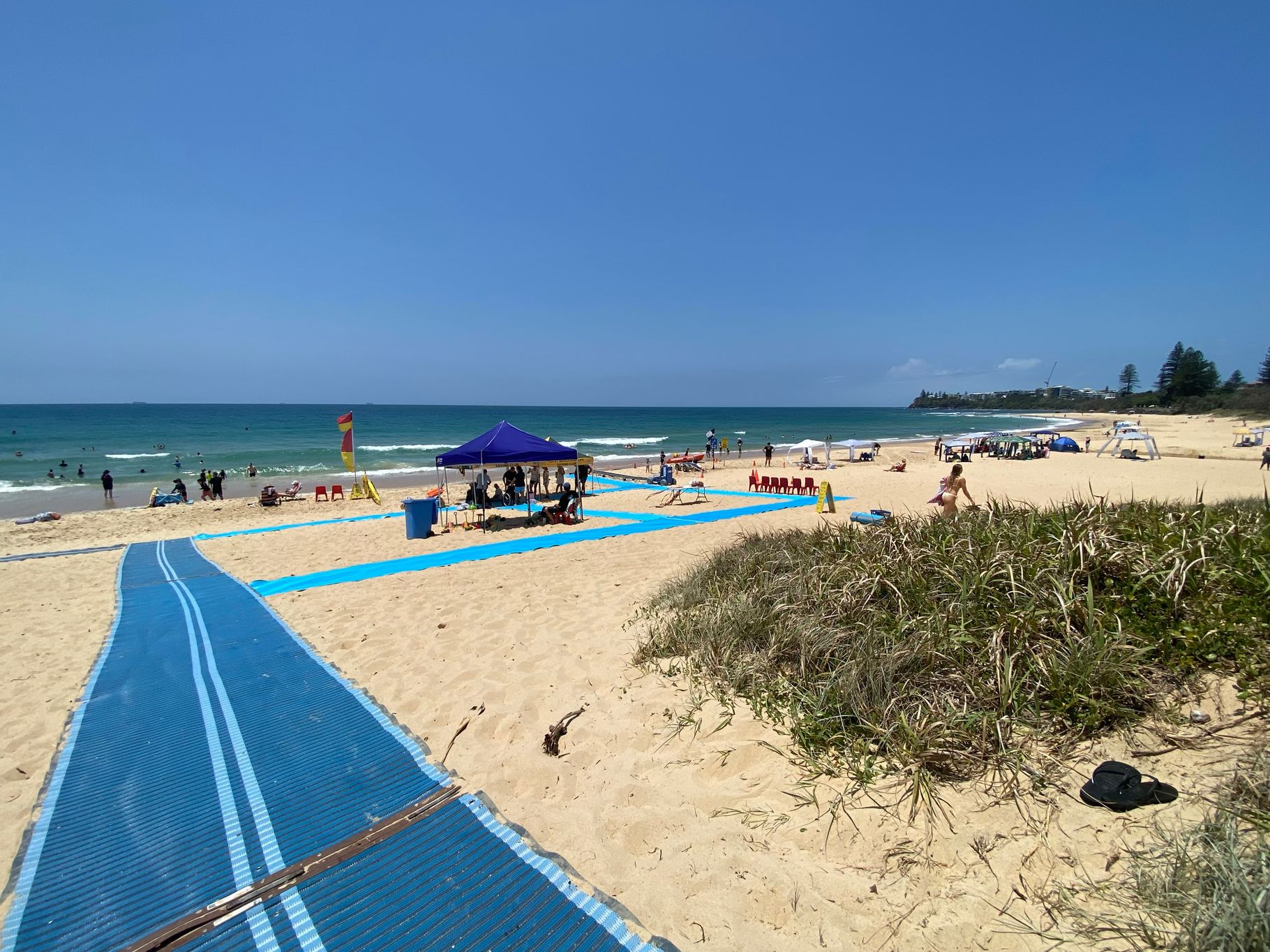 On the left, a blue mobility mat starts near the edge of the image, leading towards the centre across sandy ground. On the right, a patch of grass and low shrubs borders the sand. In the middle, a larger section of the mat runs through the sand towards a purple canopy shelter with people seated underneath. The background includes the ocean, swimmers, a surf lifesaving flag, scattered beach chairs, and other shelters. The floor is sandy with areas of vegetation. On the left, a blue mobility mat starts near the edge of the image, leading towards the centre across sandy ground. On the right, a patch of grass and low shrubs borders the sand. In the middle, a larger section of the mat runs through the sand towards a purple canopy shelter with people seated underneath. The background includes the ocean, swimmers, a surf lifesaving flag, scattered beach chairs, and other shelters. The floor is sandy with areas of vegetation.