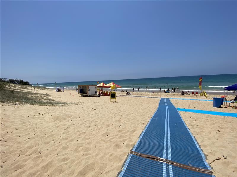 On the left, a grassy dune runs parallel to the sandy beach, with scattered vegetation at its base. On the right, part of a blue mobility mat leads to the beach, next to a blue bin and a smaller mat. In the middle, the main blue mat stretches across the sand towards a yellow and red tent near a trailer. The background includes people swimming in the ocean, a surf lifesaving flag on the right, and additional tents and chairs near the shoreline. The floor is dry sand with footprints. On the left, a grassy dune runs parallel to the sandy beach, with scattered vegetation at its base. On the right, part of a blue mobility mat leads to the beach, next to a blue bin and a smaller mat. In the middle, the main blue mat stretches across the sand towards a yellow and red tent near a trailer. The background includes people swimming in the ocean, a surf lifesaving flag on the right, and additional tents and chairs near the shoreline. The floor is dry sand with footprints.