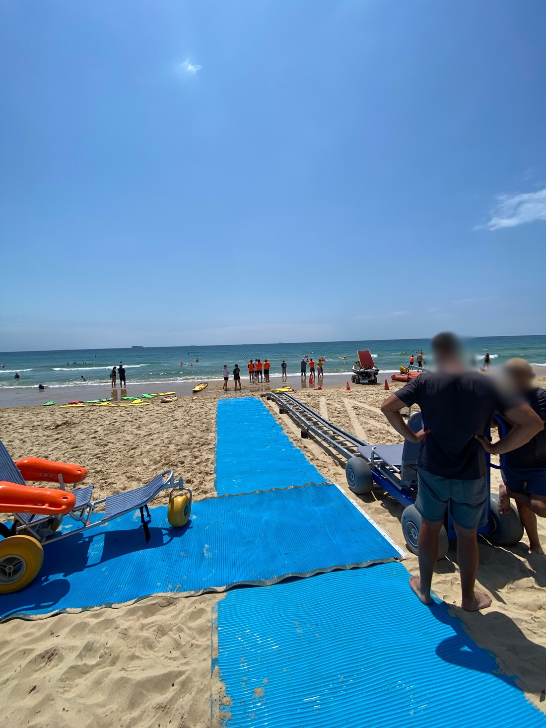 On the left, two beach wheelchairs with orange floats and large yellow wheels rest on a blue mobility mat that stretches across the sandy beach. On the right, a group of people stand near additional equipment, including another beach wheelchair with grey wheels. In the middle, the mat leads down to the shoreline where lifeguards in orange tops stand near the water. The background consists of the ocean with swimmers. The floor is sandy with footprints, and the mat has visible wear. On the left, two beach wheelchairs with orange floats and large yellow wheels rest on a blue mobility mat that stretches across the sandy beach. On the right, a group of people stand near additional equipment, including another beach wheelchair with grey wheels. In the middle, the mat leads down to the shoreline where lifeguards in orange tops stand near the water. The background consists of the ocean with swimmers. The floor is sandy with footprints, and the mat has visible wear.