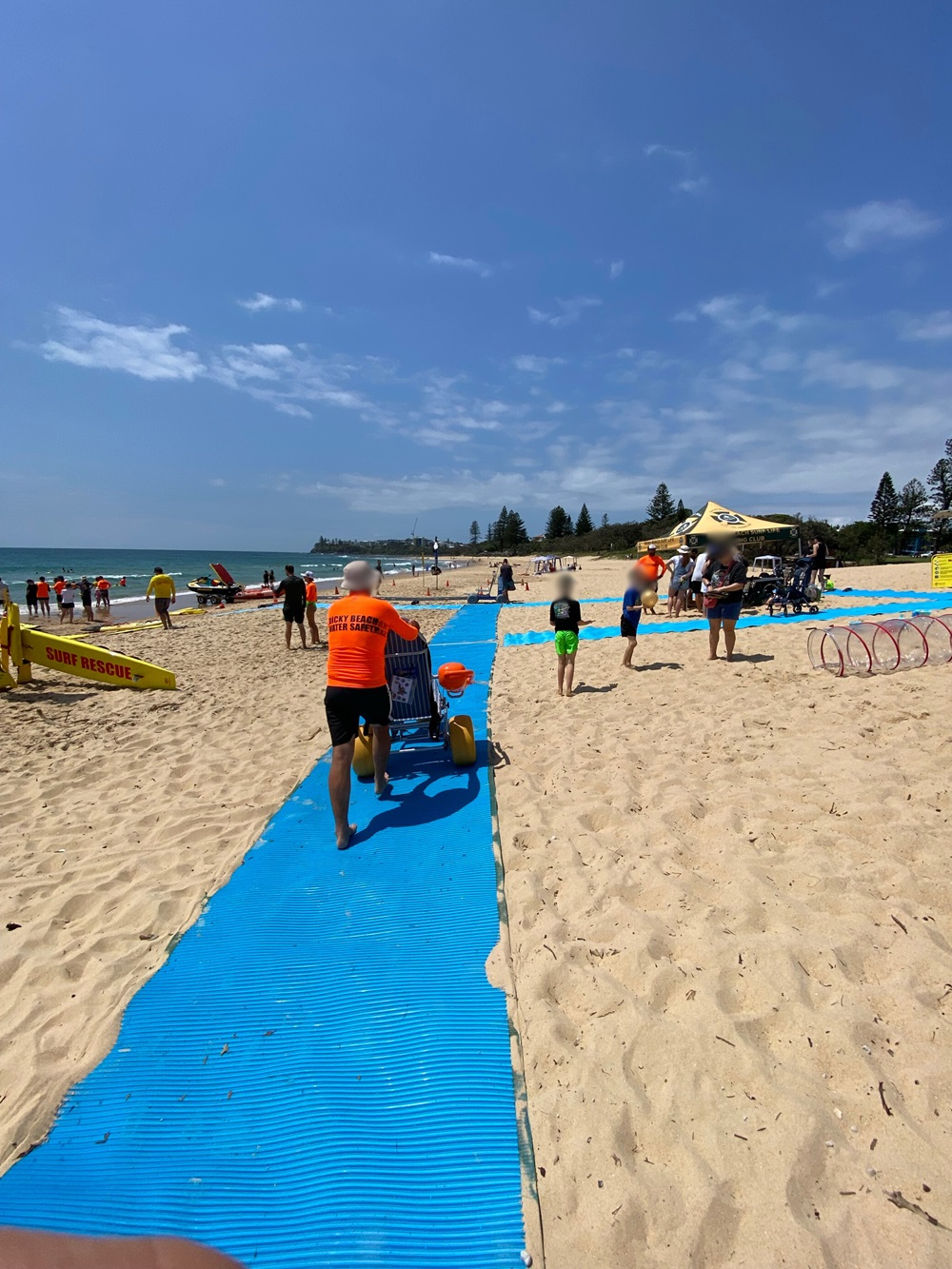 On the left, a lifeguard in an orange shirt pushes a yellow beach wheelchair along a blue mobility mat surrounded by sandy ground. On the right, a group of people gather near a yellow canopy tent featuring surf rescue equipment and hula hoops. In the middle, the mat extends straight through the sand towards the shoreline in the background, where more people stand near the water and lifeguard boats. The floor is sandy with scattered footprints, and the mat forms visible pathways. On the left, a lifeguard in an orange shirt pushes a yellow beach wheelchair along a blue mobility mat surrounded by sandy ground. On the right, a group of people gather near a yellow canopy tent featuring surf rescue equipment and hula hoops. In the middle, the mat extends straight through the sand towards the shoreline in the background, where more people stand near the water and lifeguard boats. The floor is sandy with scattered footprints, and the mat forms visible pathways.