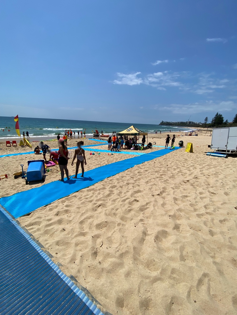 On the left, a blue mobility mat stretches over sandy ground, bordered by a lifeguard flag and beach chairs near the shoreline. On the right, a yellow canopy tent shelters people, with a white trailer and a warning sign further back on the sand. In the middle, another section of the mat extends towards the foreground, bordered by people walking and sitting on the sand. The background includes the ocean edge and distant trees. The floor is sandy, with footprints scattered across the area. On the left, a blue mobility mat stretches over sandy ground, bordered by a lifeguard flag and beach chairs near the shoreline. On the right, a yellow canopy tent shelters people, with a white trailer and a warning sign further back on the sand. In the middle, another section of the mat extends towards the foreground, bordered by people walking and sitting on the sand. The background includes the ocean edge and distant trees. The floor is sandy, with footprints scattered across the area.
