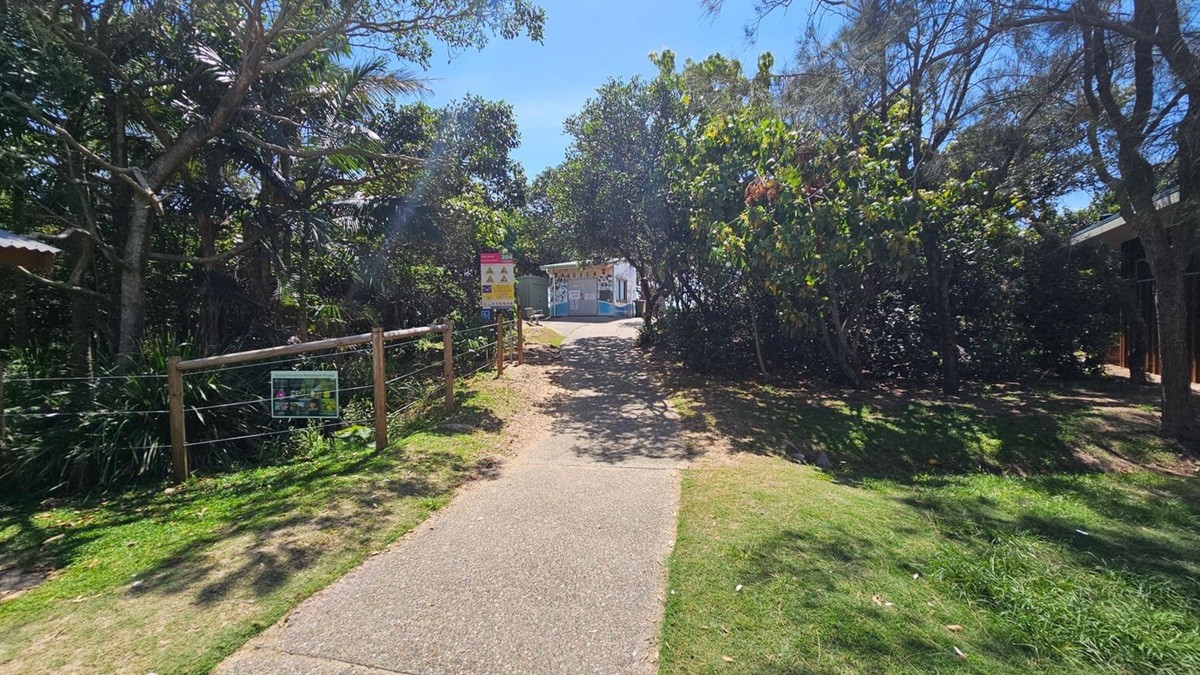 A path runs centre, flanked by grass and dense bushes. To the left, a wooden fence with wire fencing sits along the edge of a trimmed lawn; a small signboard is mounted on the fence. On the right, thick shrubbery and trees create a shaded verge. In the distance, a small building or booth is visible beyond the path, with more greenery framing the scene. The ground is a light-paved footpath; grass borders both sides. A path runs centre, flanked by grass and dense bushes. To the left, a wooden fence with wire fencing sits along the edge of a trimmed lawn; a small signboard is mounted on the fence. On the right, thick shrubbery and trees create a shaded verge. In the distance, a small building or booth is visible beyond the path, with more greenery framing the scene. The ground is a light-paved footpath; grass borders both sides.