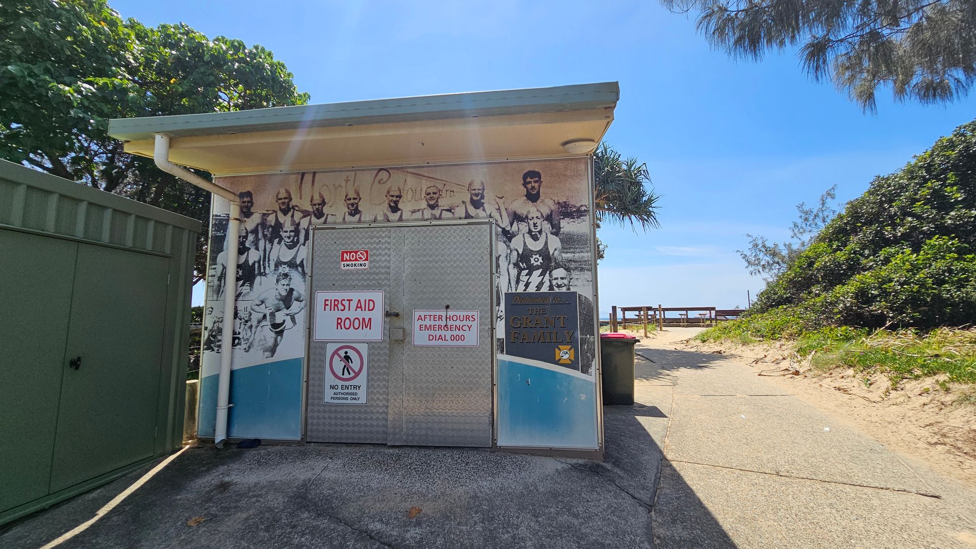 A small structure with a bold metal door in the centre. Left side shows a green utility cabinet and a white pipe running down the wall; right side has a brown rubbish bin. Background features a wooden railing path leading to a sandy slope with bushes on either side. The floor is a grey concrete surface, slightly worn, extending from the front of the door to the path on the right. Ceiling is a flat, light-coloured canopy. A small structure with a bold metal door in the centre. Left side shows a green utility cabinet and a white pipe running down the wall; right side has a brown rubbish bin. Background features a wooden railing path leading to a sandy slope with bushes on either side. The floor is a grey concrete surface, slightly worn, extending from the front of the door to the path on the right. Ceiling is a flat, light-coloured canopy.