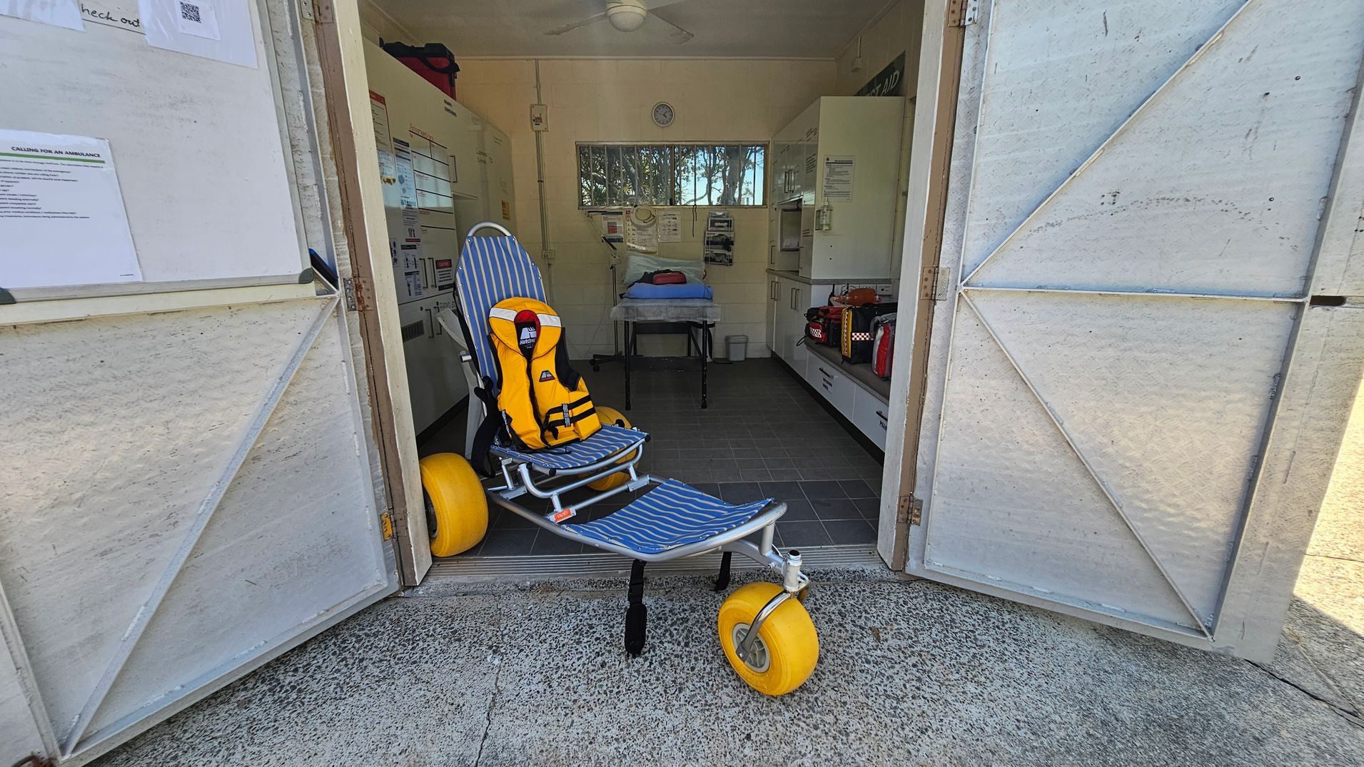 A doorway opens into a small medical room. On the left inside, a tall metal cabinet lines the wall. Center is a blue-and-white striped reclining chair with a bright yellow life jacket secured on it. A single large yellow wheel sits at the chair’s rear. In the middle/background, a hospital trolley with a blue sheet stands near a workbench and white cupboards. The floor is dark grey tiles inside; the outer threshold is rough concrete. A doorway opens into a small medical room. On the left inside, a tall metal cabinet lines the wall. Center is a blue-and-white striped reclining chair with a bright yellow life jacket secured on it. A single large yellow wheel sits at the chair’s rear. In the middle/background, a hospital trolley with a blue sheet stands near a workbench and white cupboards. The floor is dark grey tiles inside; the outer threshold is rough concrete.