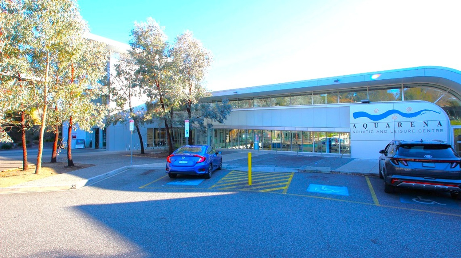 A modern building with a curved roof on the right, featuring large windows and a sign reading "Aquarena Aquatic and Leisure Centre." Two cars are parked in front, one blue and one black, with a disabled parking space marked on the ground. On the left, several tall trees line the pavement, casting shadows on the ground. The area is well-lit, and the road in the foreground is smooth and wide. A modern building with a curved roof on the right, featuring large windows and a sign reading "Aquarena Aquatic and Leisure Centre." Two cars are parked in front, one blue and one black, with a disabled parking space marked on the ground. On the left, several tall trees line the pavement, casting shadows on the ground. The area is well-lit, and the road in the foreground is smooth and wide.