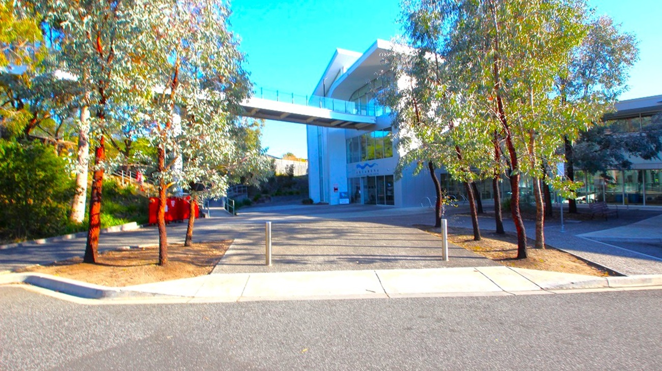 A modern building with a white bridge connecting two sections is in the centre. Trees with slender trunks and sparse foliage are on both sides, casting shadows on the ground. The foreground features a paved area with bollards and a kerb, leading to a road. The building has large glass windows, and the area is well-lit, suggesting a sunny day. A modern building with a white bridge connecting two sections is in the centre. Trees with slender trunks and sparse foliage are on both sides, casting shadows on the ground. The foreground features a paved area with bollards and a kerb, leading to a road. The building has large glass windows, and the area is well-lit, suggesting a sunny day.