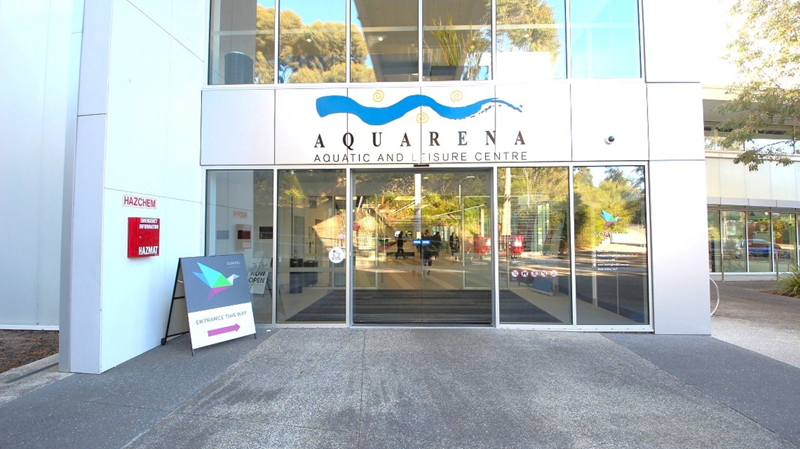 Entrance to a building with large glass doors and windows. Above the doors, a sign reads "Aquarena Aquatic and Leisure Centre" with a wave design. To the left, a red sign indicates "Hazchem" and "Mazmat," and a sandwich board sign is placed on the ground. The floor is paved, and reflections of trees and the surroundings are visible in the glass. Trees and greenery are seen in the background through the glass. Entrance to a building with large glass doors and windows. Above the doors, a sign reads "Aquarena Aquatic and Leisure Centre" with a wave design. To the left, a red sign indicates "Hazchem" and "Mazmat," and a sandwich board sign is placed on the ground. The floor is paved, and reflections of trees and the surroundings are visible in the glass. Trees and greenery are seen in the background through the glass.