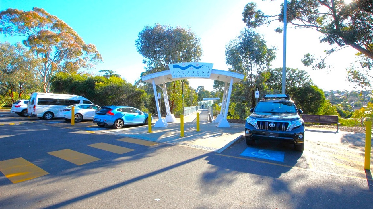 A parking area with several vehicles, including a white van and a blue car on the left, and a black SUV on the right. In the centre, a white archway with a sign reading "Quarry" leads to a pathway. Yellow bollards are in front of the archway. Trees and greenery surround the area, with a bench visible on the right. The ground is marked with yellow pedestrian lines, and a tall streetlight is on the right. A parking area with several vehicles, including a white van and a blue car on the left, and a black SUV on the right. In the centre, a white archway with a sign reading "Quarry" leads to a pathway. Yellow bollards are in front of the archway. Trees and greenery surround the area, with a bench visible on the right. The ground is marked with yellow pedestrian lines, and a tall streetlight is on the right.