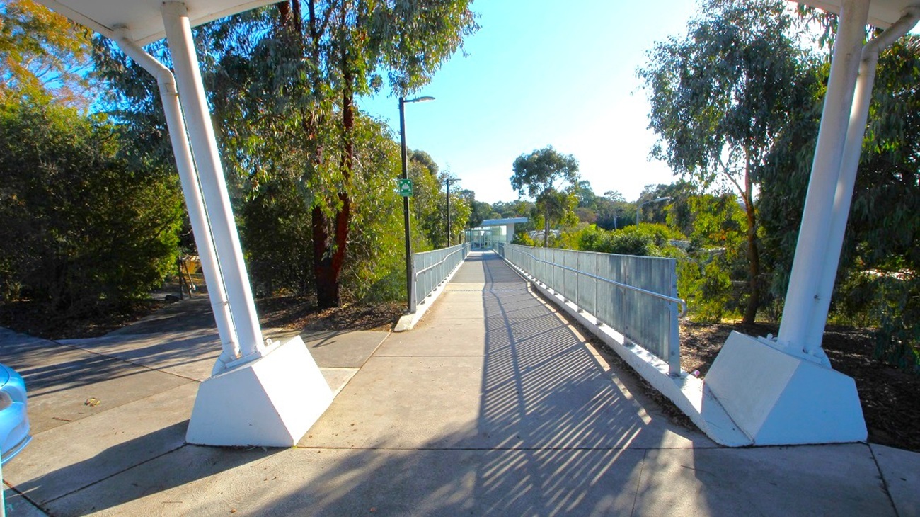 A walkway with a metal railing on both sides extends into the distance, flanked by tall trees on the left and right. The path is paved and slightly elevated, with shadows cast by the railings. In the foreground, there are two large white support structures on either side. A blue car is partially visible on the left. The background features more trees and a clear sky. A walkway with a metal railing on both sides extends into the distance, flanked by tall trees on the left and right. The path is paved and slightly elevated, with shadows cast by the railings. In the foreground, there are two large white support structures on either side. A blue car is partially visible on the left. The background features more trees and a clear sky.