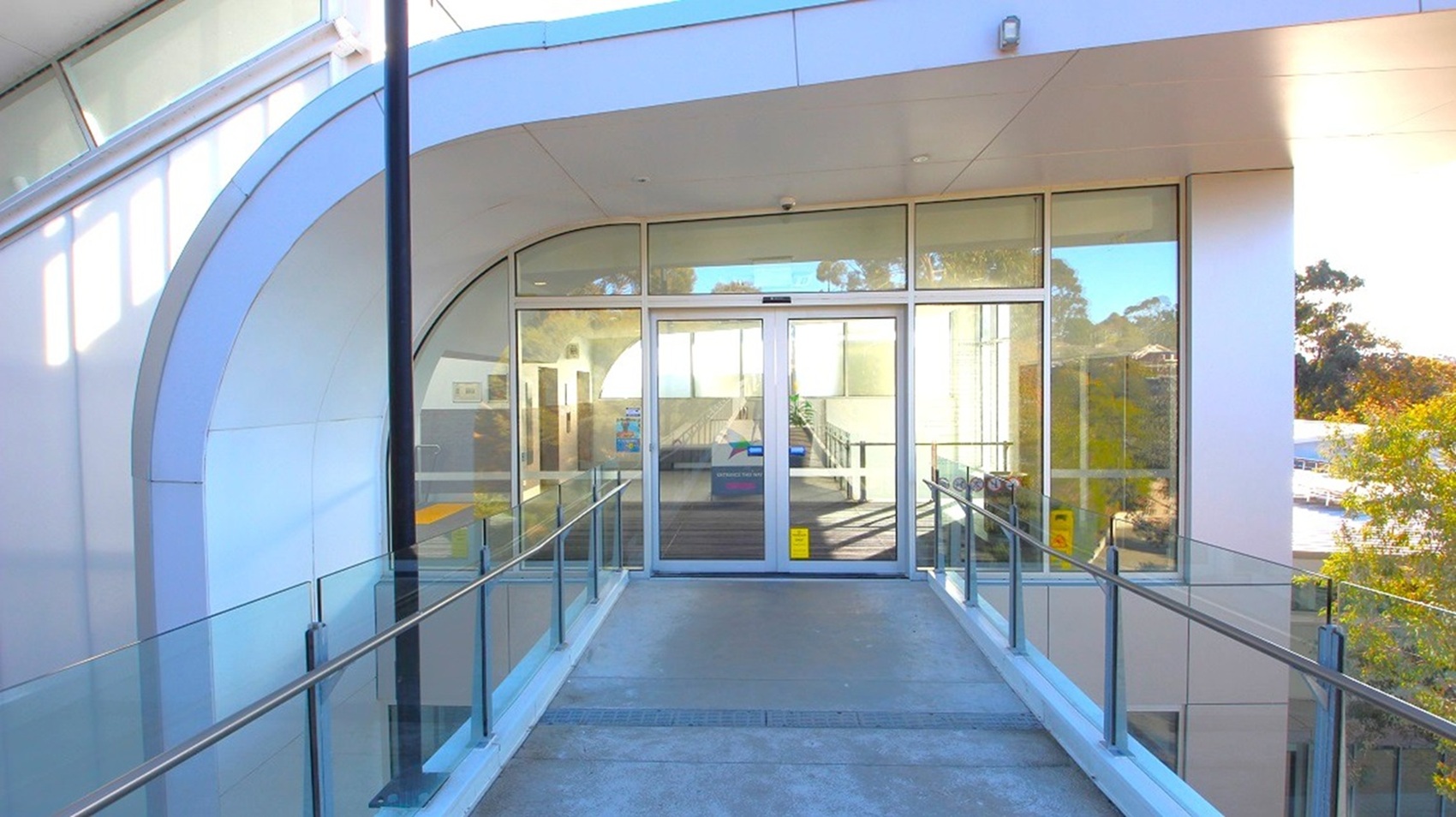 A modern entrance with glass double doors in the centre, surrounded by large glass panels. The structure has a curved design with a white exterior. A concrete ramp with metal railings leads up to the doors. On the right, trees are visible through the glass. The ceiling is smooth and white, with a light fixture above the doors. A modern entrance with glass double doors in the centre, surrounded by large glass panels. The structure has a curved design with a white exterior. A concrete ramp with metal railings leads up to the doors. On the right, trees are visible through the glass. The ceiling is smooth and white, with a light fixture above the doors.