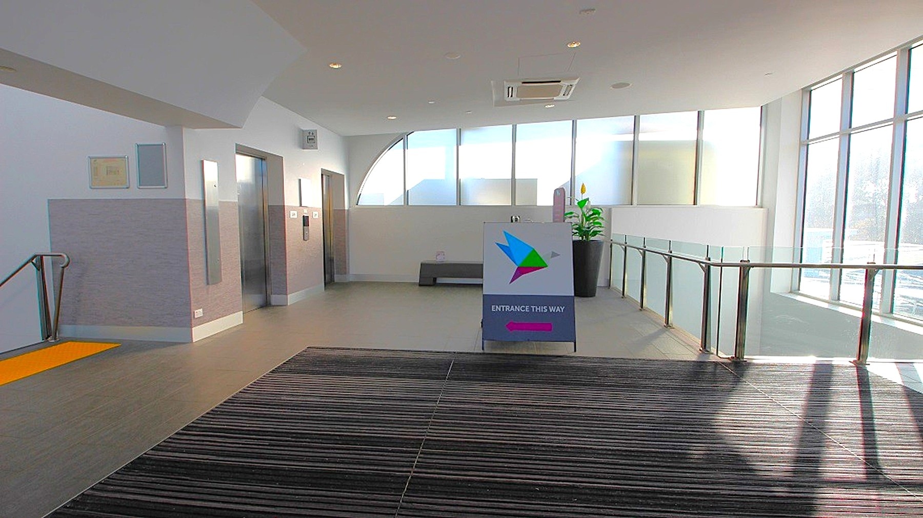 A modern indoor space with a striped carpeted floor in the foreground. On the left, there are two lift doors and a staircase with a yellow safety strip. In the centre, a sign with a colourful logo and text directs to an entrance. The right side features large windows allowing natural light to flood in, with a glass railing and a potted plant nearby. The ceiling is white with recessed lighting and an air conditioning unit. A modern indoor space with a striped carpeted floor in the foreground. On the left, there are two lift doors and a staircase with a yellow safety strip. In the centre, a sign with a colourful logo and text directs to an entrance. The right side features large windows allowing natural light to flood in, with a glass railing and a potted plant nearby. The ceiling is white with recessed lighting and an air conditioning unit.