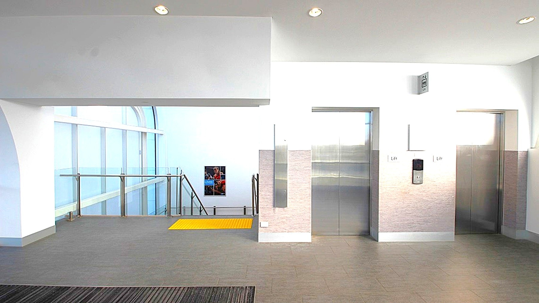 A modern interior space with two lift doors on the right, featuring metallic finishes. To the left, a staircase with a glass railing descends, and a yellow safety strip is visible on the floor near the stairs. The walls are white, and the ceiling has recessed lighting. The floor is grey, complementing the minimalist design. A small window is visible in the background, providing natural light. A modern interior space with two lift doors on the right, featuring metallic finishes. To the left, a staircase with a glass railing descends, and a yellow safety strip is visible on the floor near the stairs. The walls are white, and the ceiling has recessed lighting. The floor is grey, complementing the minimalist design. A small window is visible in the background, providing natural light.
