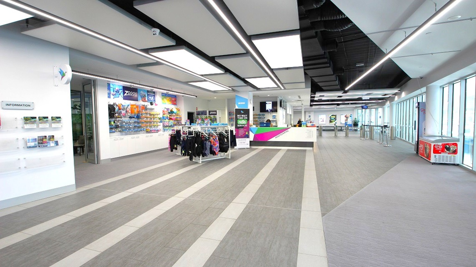 A spacious, modern interior with a grey-striped floor and a ceiling featuring rectangular light panels. On the left, there is a wall with information displays and a shop area with racks of clothing and shelves of merchandise. In the centre, a digital sign stands near a counter. The right side has large windows allowing natural light, and a small vending machine is visible. The area is open and well-lit, with a sleek, contemporary design. A spacious, modern interior with a grey-striped floor and a ceiling featuring rectangular light panels. On the left, there is a wall with information displays and a shop area with racks of clothing and shelves of merchandise. In the centre, a digital sign stands near a counter. The right side has large windows allowing natural light, and a small vending machine is visible. The area is open and well-lit, with a sleek, contemporary design.