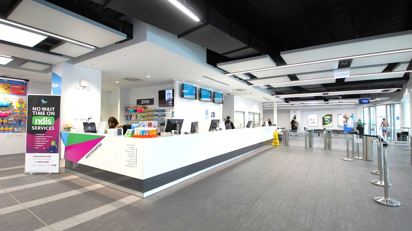 A modern interior with a long white reception desk on the right, equipped with computers and attended by staff. Above the desk are several digital screens. On the left, a colourful banner advertises "NDIS Services." The floor is grey with a tiled pattern, and the ceiling features a grid of black and white panels with integrated lighting. In the background, there are posters on the walls and a glass entrance with people entering or exiting. Queue barriers are set up in front of the desk. A modern interior with a long white reception desk on the right, equipped with computers and attended by staff. Above the desk are several digital screens. On the left, a colourful banner advertises "NDIS Services." The floor is grey with a tiled pattern, and the ceiling features a grid of black and white panels with integrated lighting. In the background, there are posters on the walls and a glass entrance with people entering or exiting. Queue barriers are set up in front of the desk.