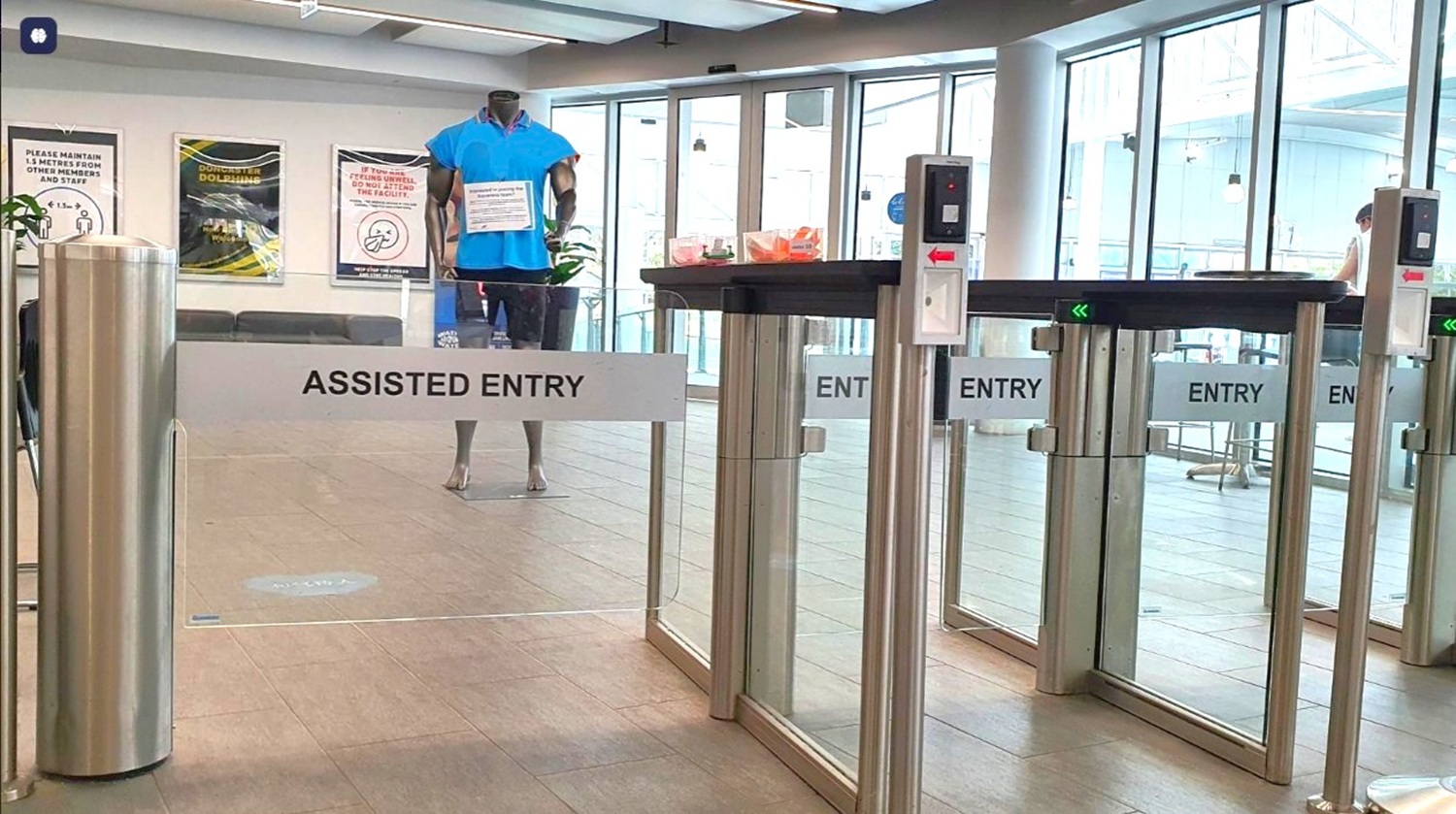 A modern entry security checkpoint with glass doors labeled “ENTRY” and a barrier marked “ASSISTED ENTRY.” A mannequin or person stands inside the entrance area, and turnstiles/automatic doors are visible. A modern entry security checkpoint with glass doors labeled “ENTRY” and a barrier marked “ASSISTED ENTRY.” A mannequin or person stands inside the entrance area, and turnstiles/automatic doors are visible.