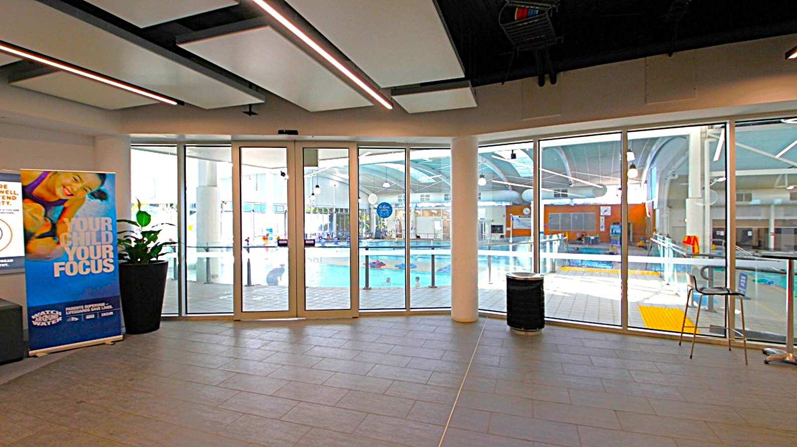 A spacious indoor area with large glass doors and windows in the centre, providing a view of a swimming pool area with clear water and poolside equipment. On the left, there's a promotional banner next to a potted plant. On the right, a tall chair and a small table are positioned near the windows. The ceiling features modern lighting fixtures, and the floor is tiled in a dark colour. A spacious indoor area with large glass doors and windows in the centre, providing a view of a swimming pool area with clear water and poolside equipment. On the left, there's a promotional banner next to a potted plant. On the right, a tall chair and a small table are positioned near the windows. The ceiling features modern lighting fixtures, and the floor is tiled in a dark colour.