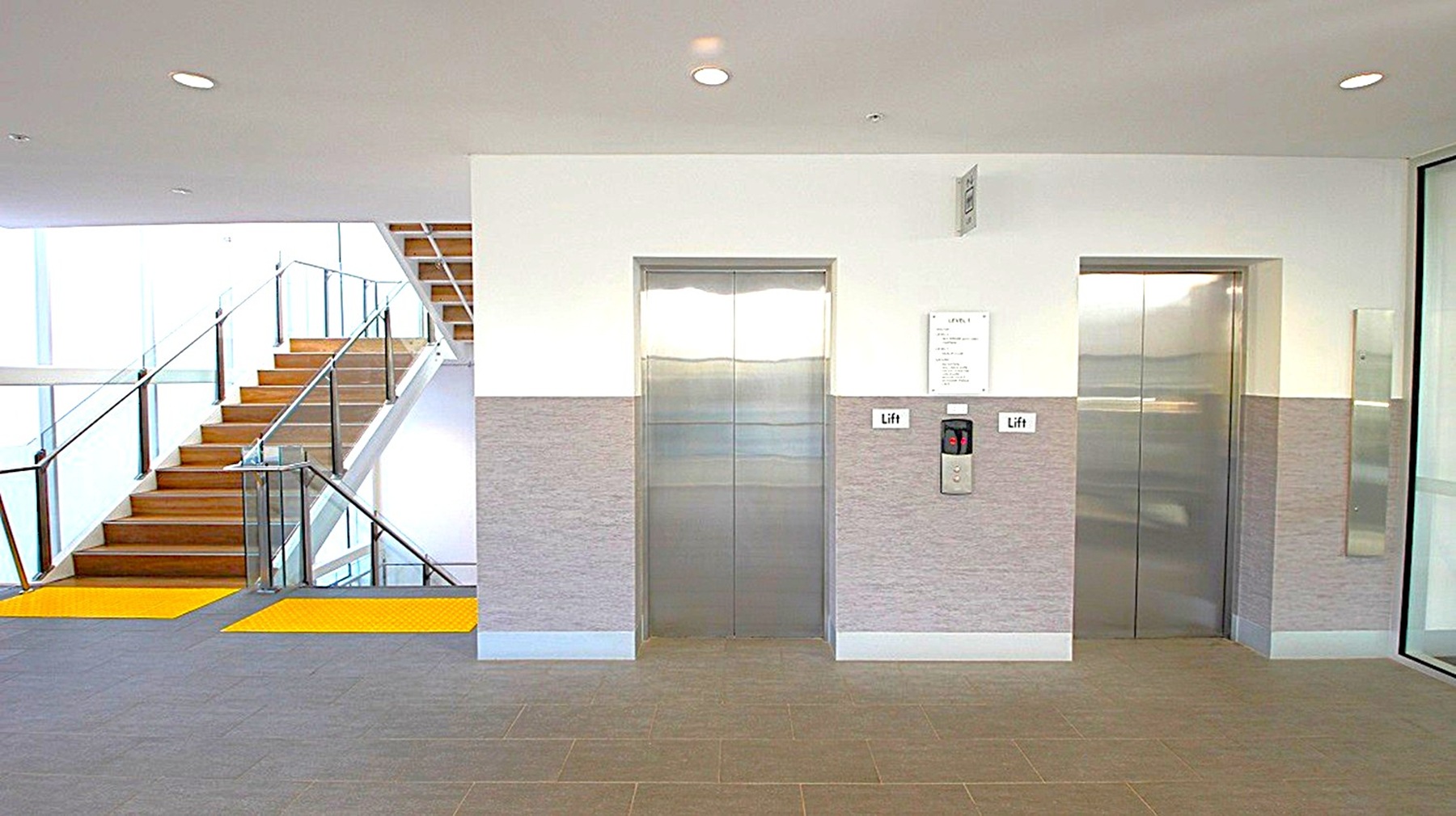 On the left, there is a staircase with wooden steps and glass railings, leading upwards. The middle features two lift doors with metallic finishes, set in a wall with a grey lower half and white upper half. A control panel and a sign labelled "Lift" are visible between the doors. On the right, a glass wall or door is partially visible. The floor is carpeted in a neutral tone, and the ceiling is white with recessed lighting. On the left, there is a staircase with wooden steps and glass railings, leading upwards. The middle features two lift doors with metallic finishes, set in a wall with a grey lower half and white upper half. A control panel and a sign labelled "Lift" are visible between the doors. On the right, a glass wall or door is partially visible. The floor is carpeted in a neutral tone, and the ceiling is white with recessed lighting.
