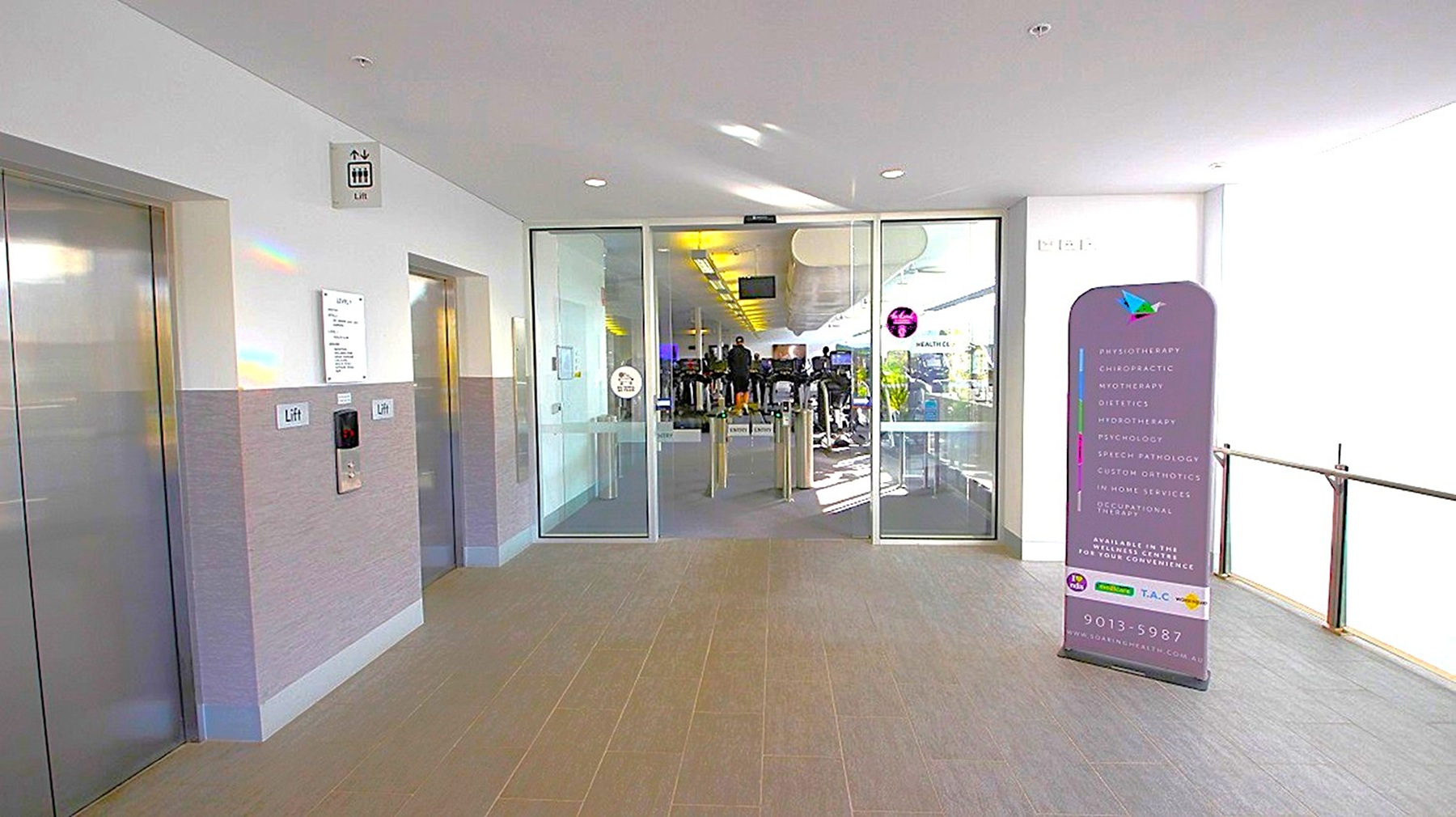 On the left, there are two lift doors with a sign above indicating their location. In the centre, glass doors lead into a gym area with exercise equipment visible inside. On the right, a purple sign with text stands near the entrance. The floor is tiled, and the ceiling is white with recessed lighting. On the left, there are two lift doors with a sign above indicating their location. In the centre, glass doors lead into a gym area with exercise equipment visible inside. On the right, a purple sign with text stands near the entrance. The floor is tiled, and the ceiling is white with recessed lighting.